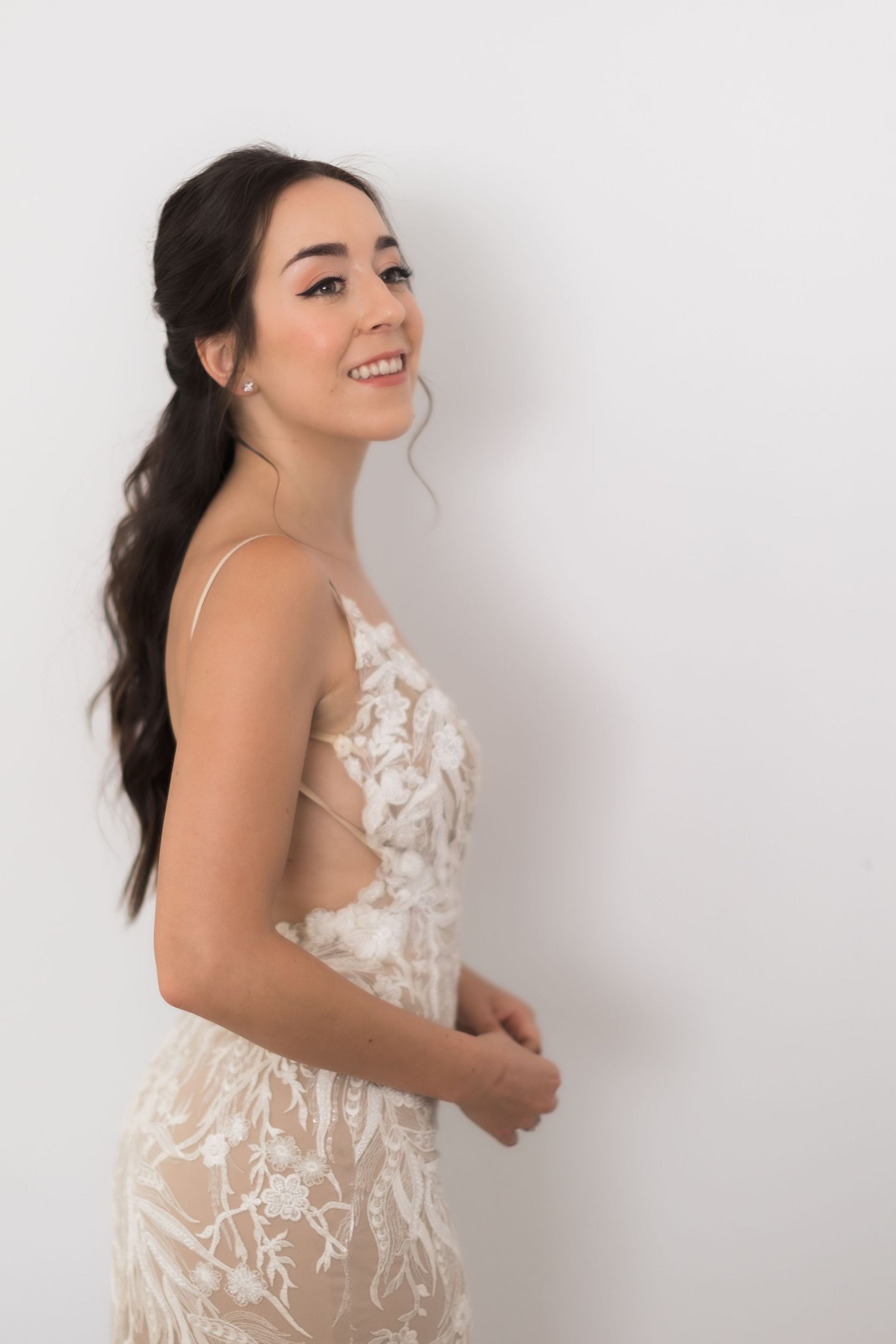 A bride poses in a sheer white lace gown with floral appliques against a white background.