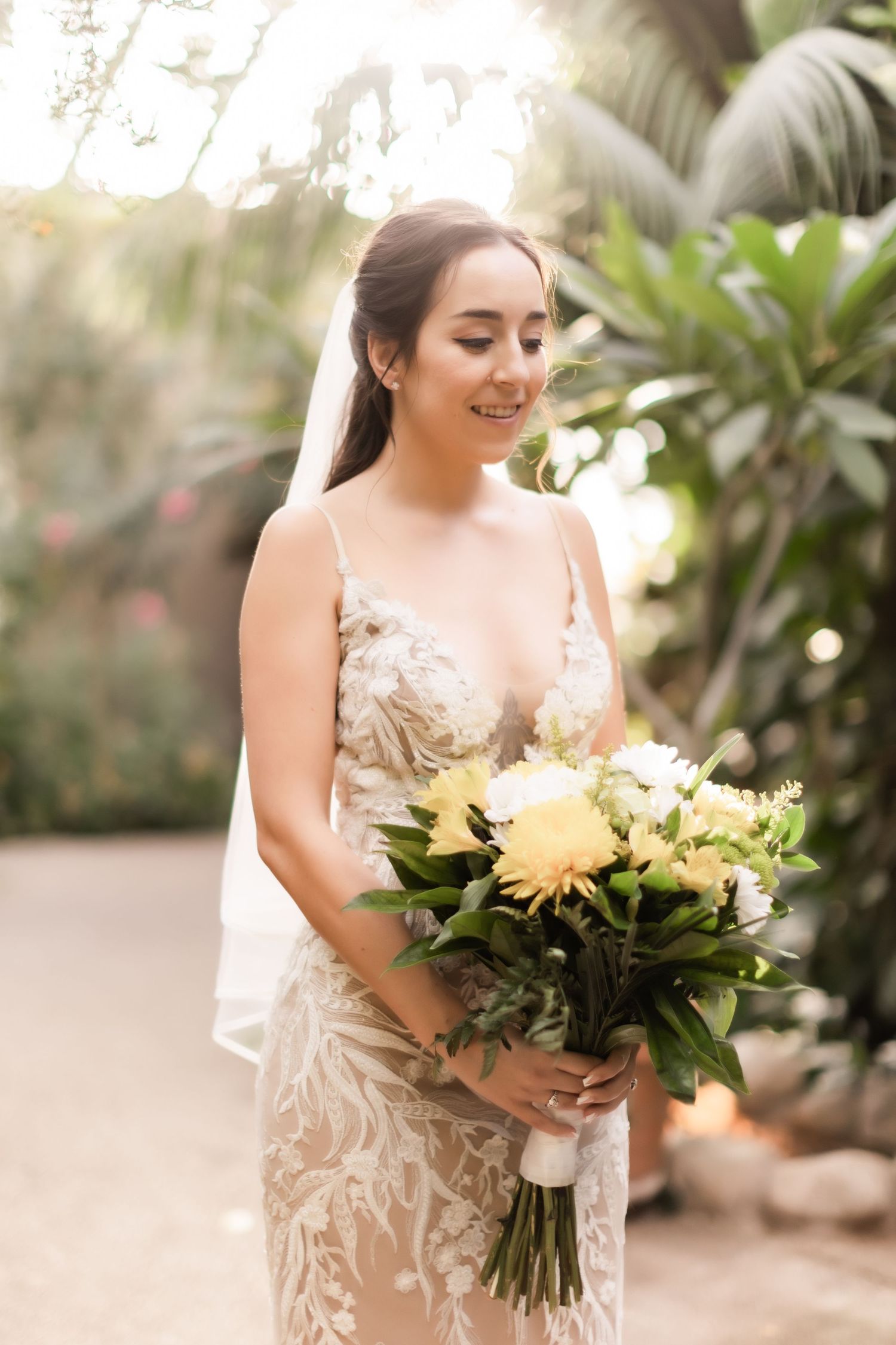 Bride holding yellow and white bouquet poses in sunlit garden setting wearing lace gown.