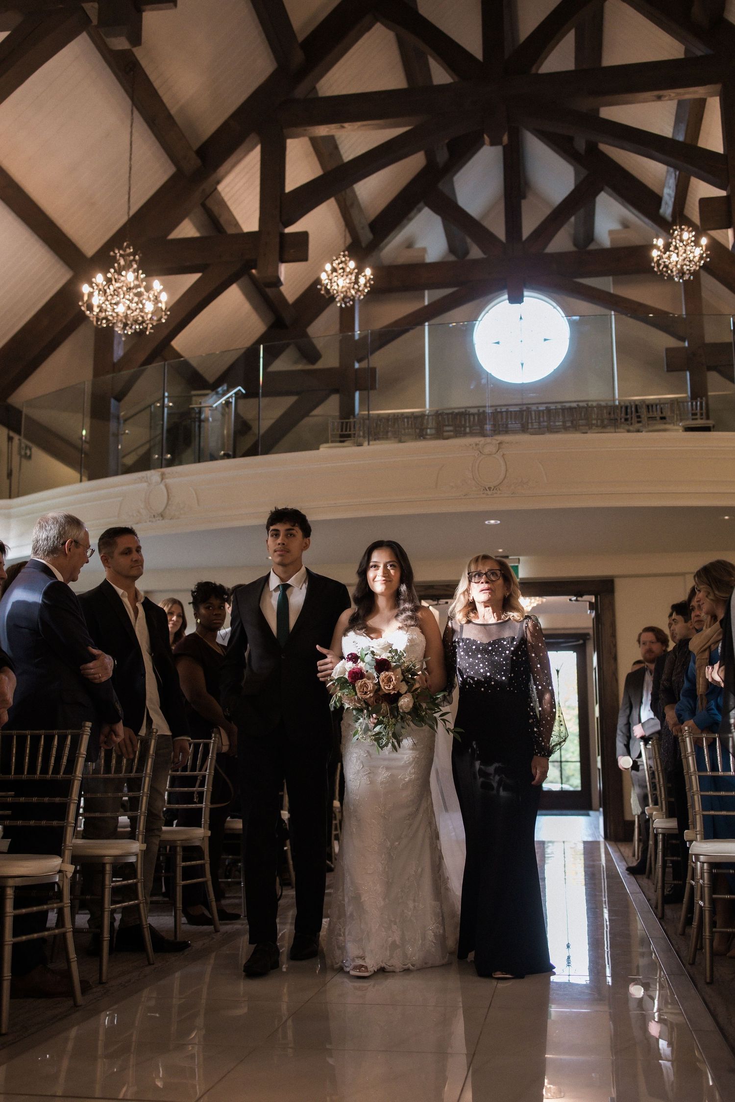 Wedding processional walking down the aisle in an elegant chapel with wooden beams and chandeliers.