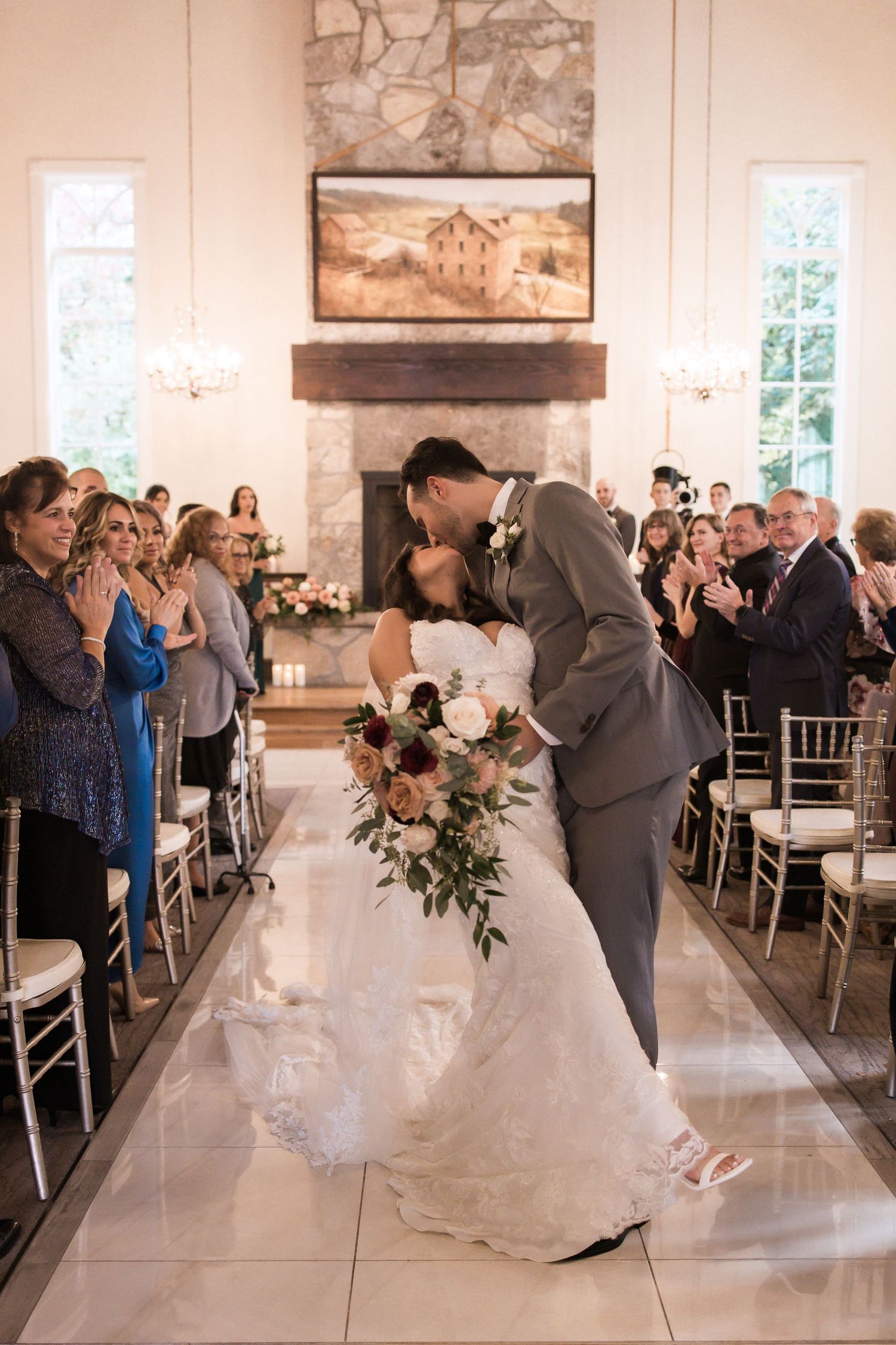 Romantic wedding kiss at ceremony with guests applauding in elegant white venue.