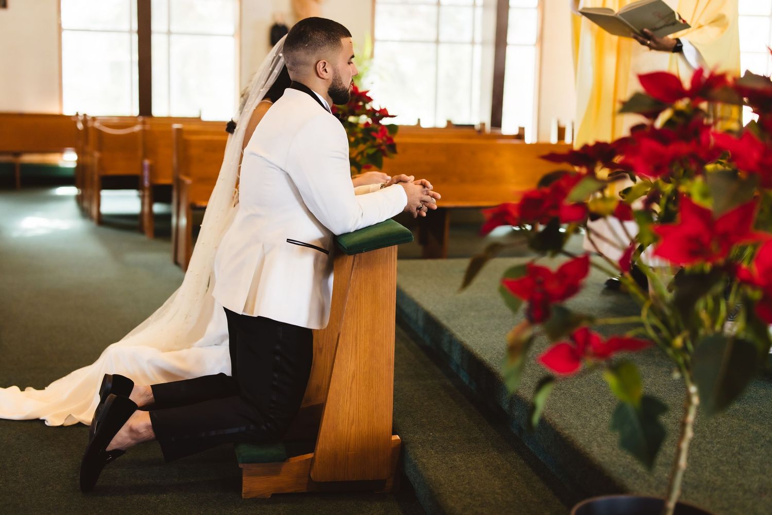 A person kneels in prayer at a church pew beside vibrant red poinsettias and tall windows.