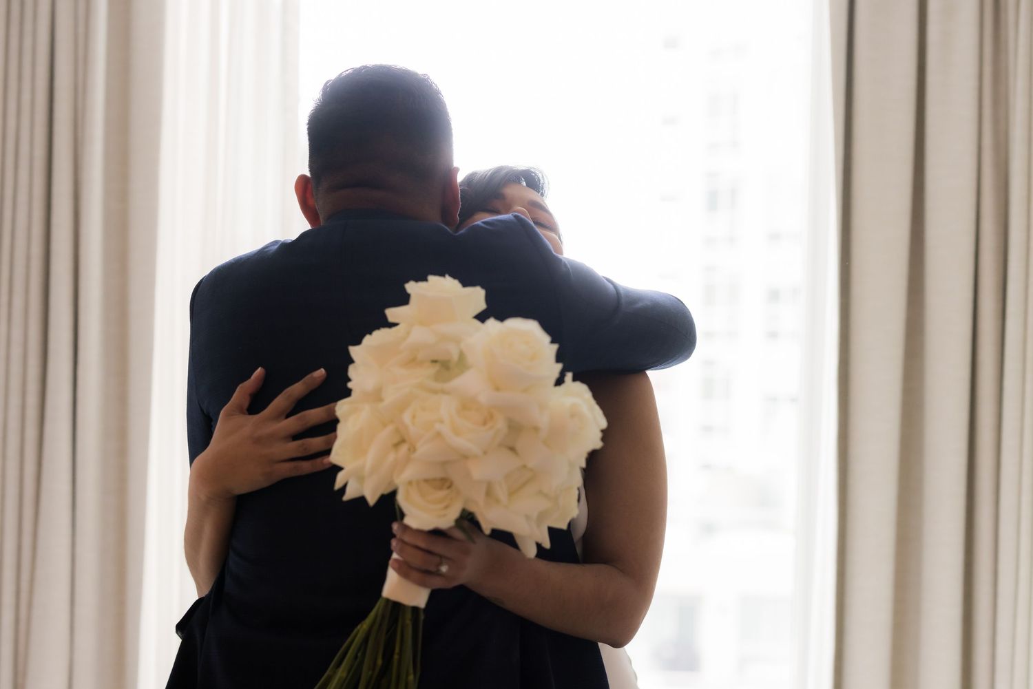 Wedding couple embraces while holding white flower bouquet in sunlit room.