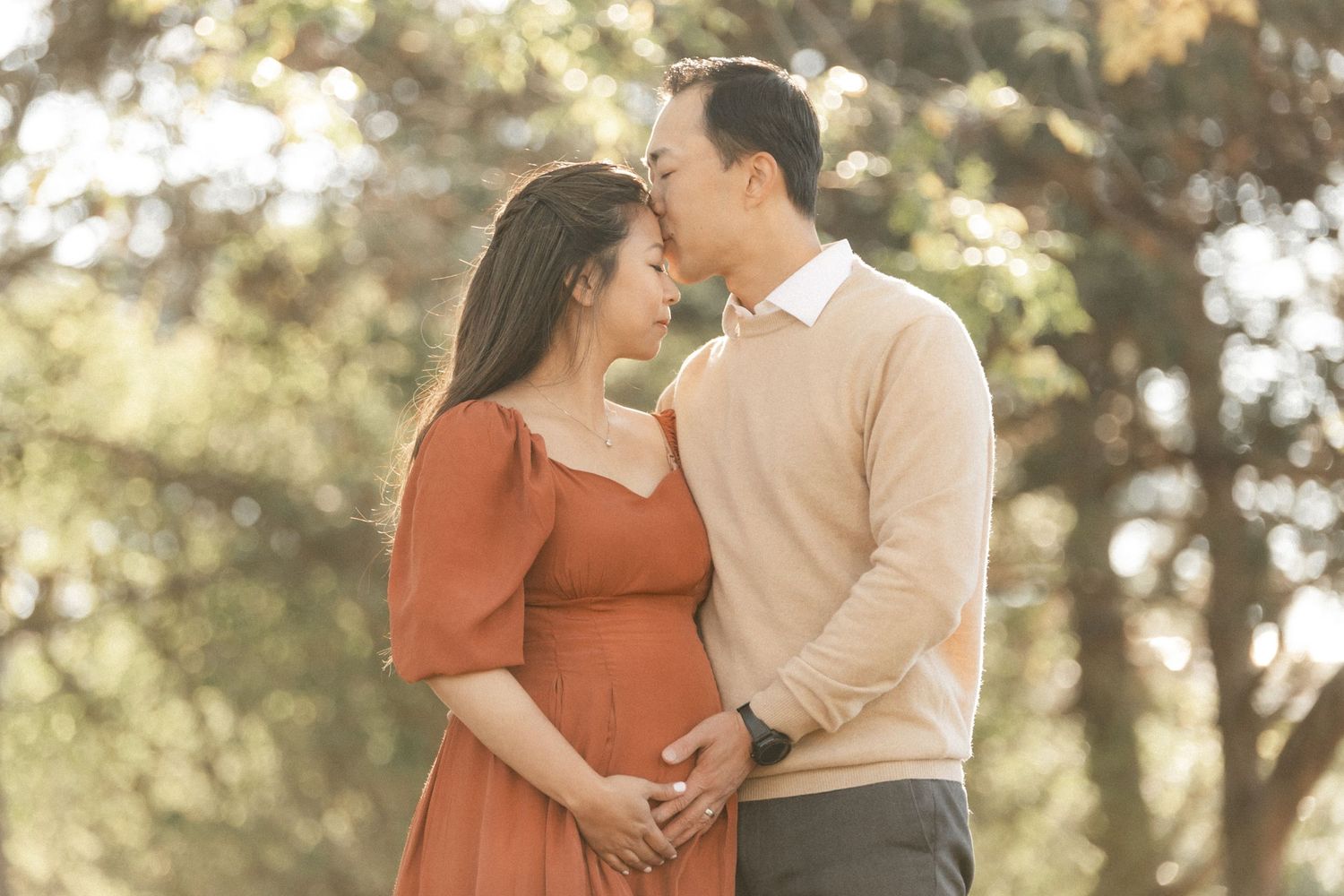 An expectant couple shares a romantic moment in a sunlit outdoor setting wearing matching earth tones.