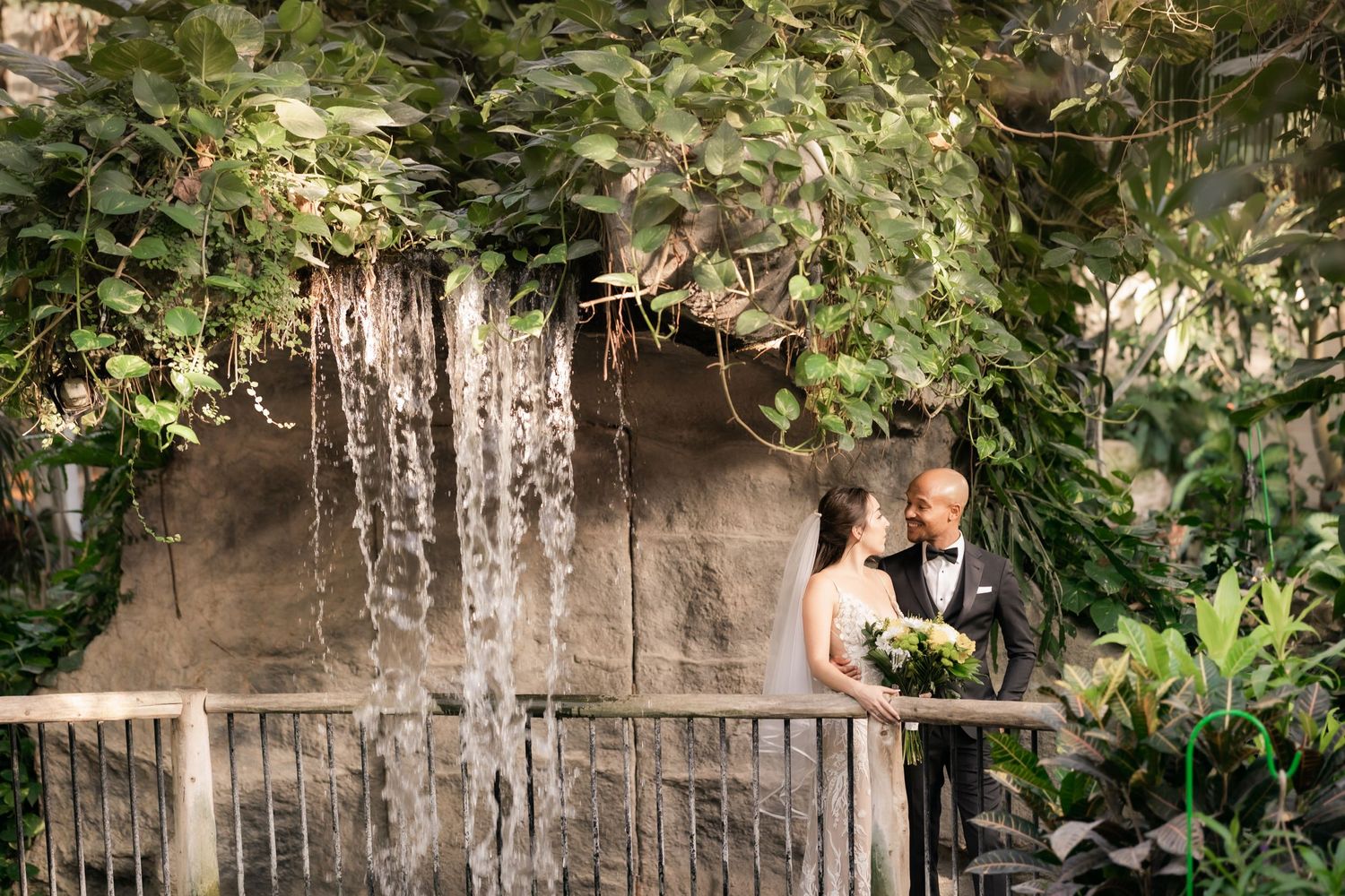 Wedding couple poses on balcony overlooking waterfall feature in botanical garden setting.