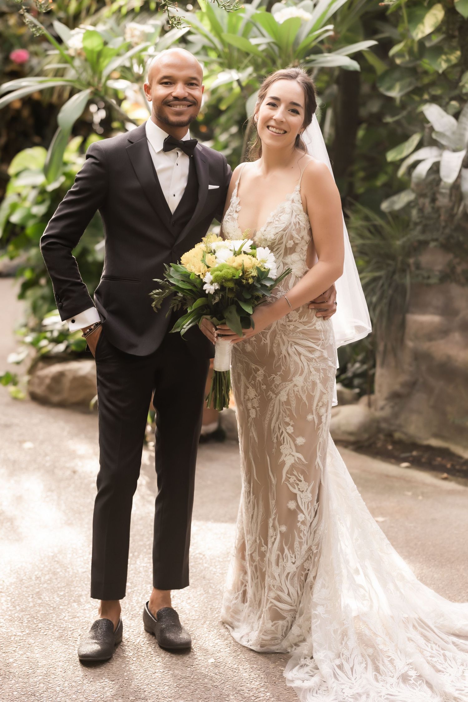 Joyful portrait of a couple in elegant wedding attire standing in a tropical garden setting.