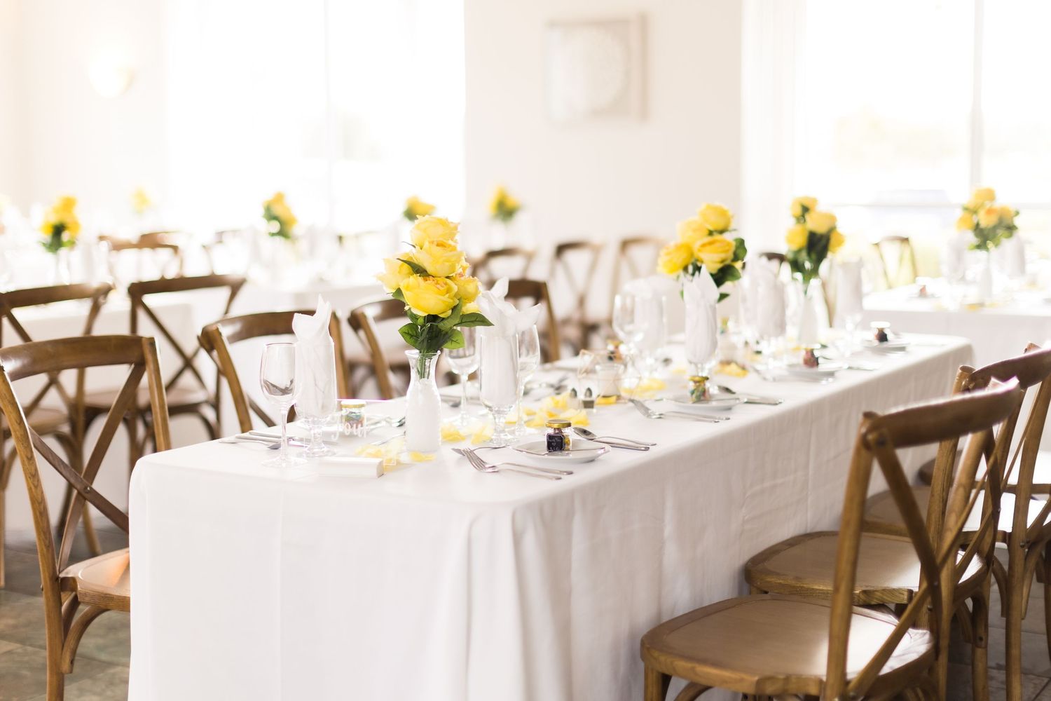 Long white dining table set with yellow flower centerpieces and wooden cross-back chairs.