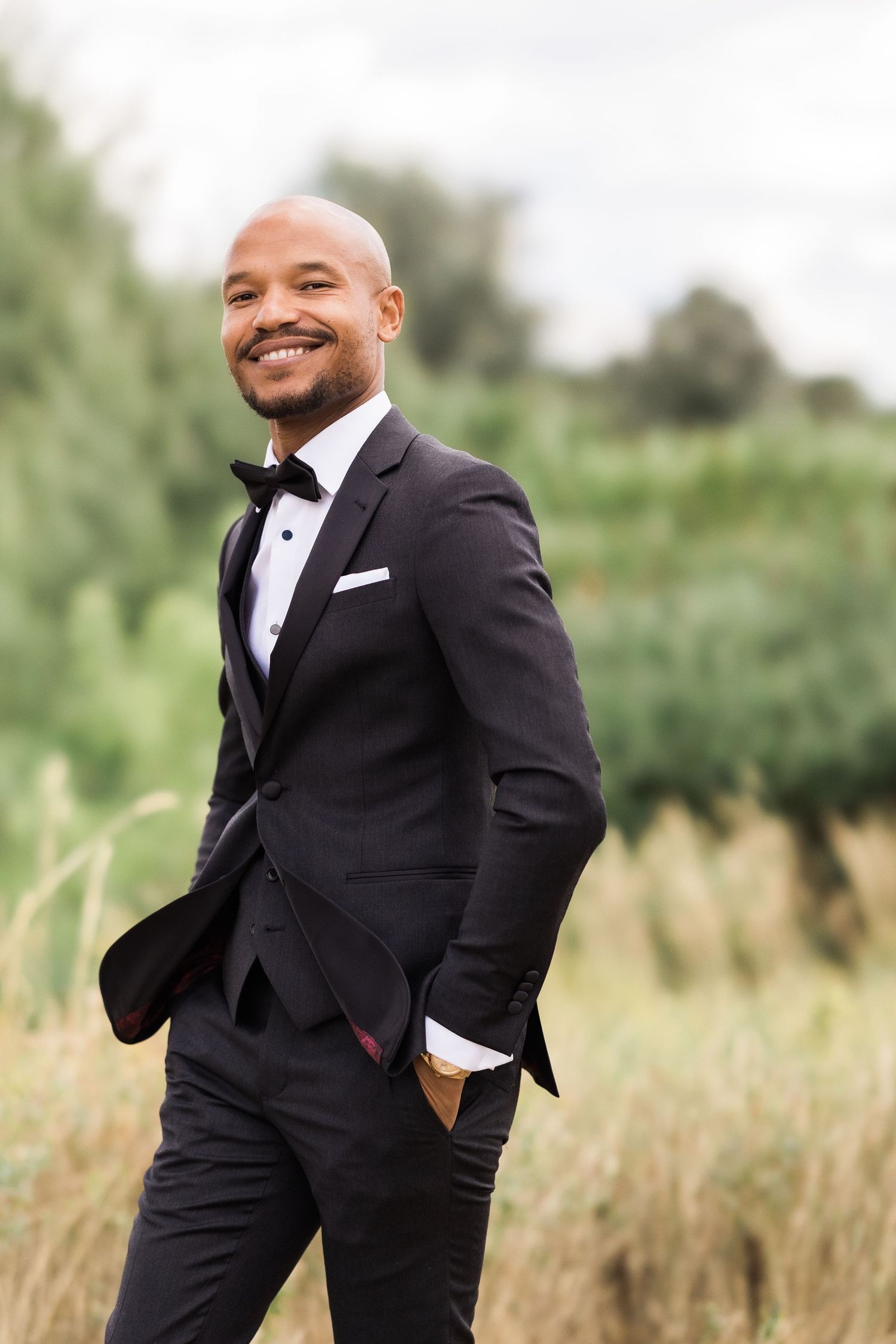 Elegant outdoor portrait of person in fitted black tuxedo posing confidently against natural backdrop.
