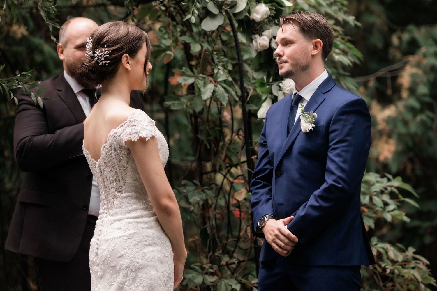 A wedding ceremony takes place in a lush garden setting with an officiant and couple in formal attire.