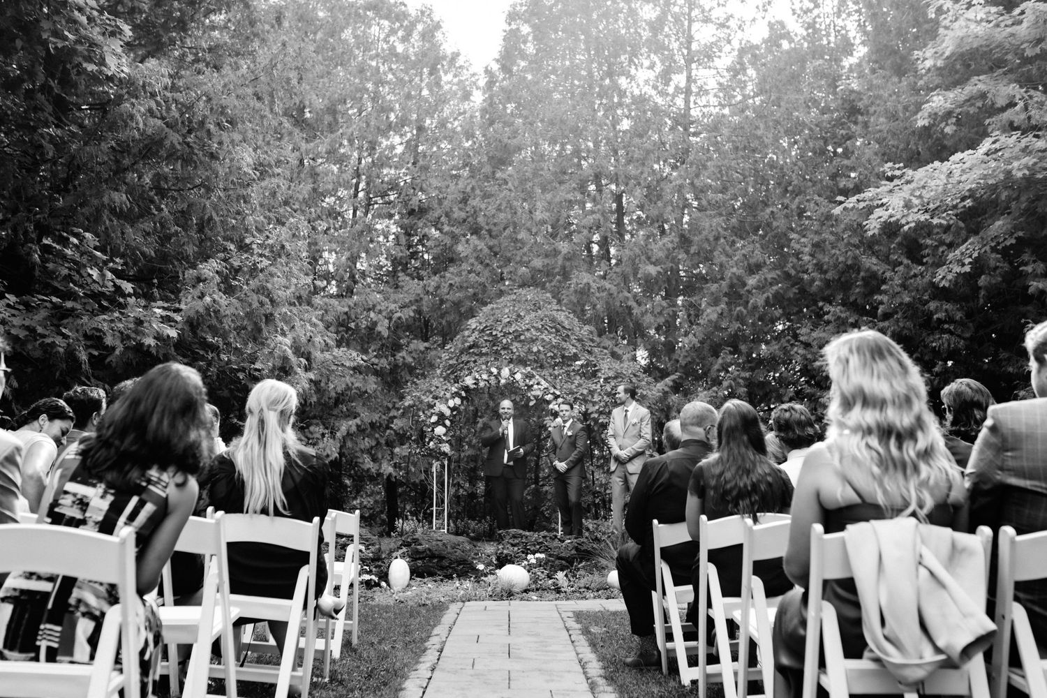 Black and white photo of outdoor forest wedding ceremony with guests seated in white chairs.