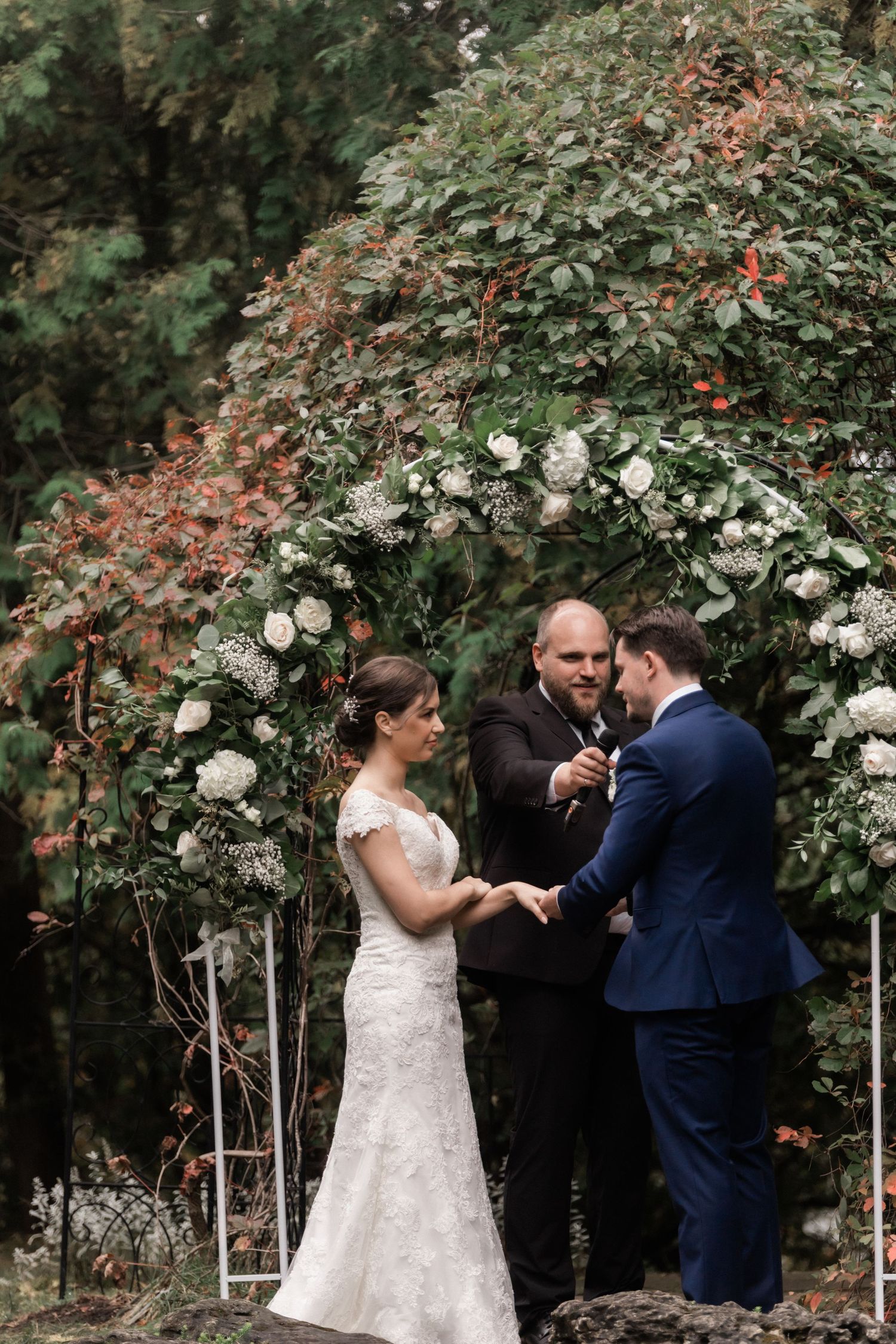 Wedding ceremony takes place beneath a beautiful floral arch with white roses and greenery.