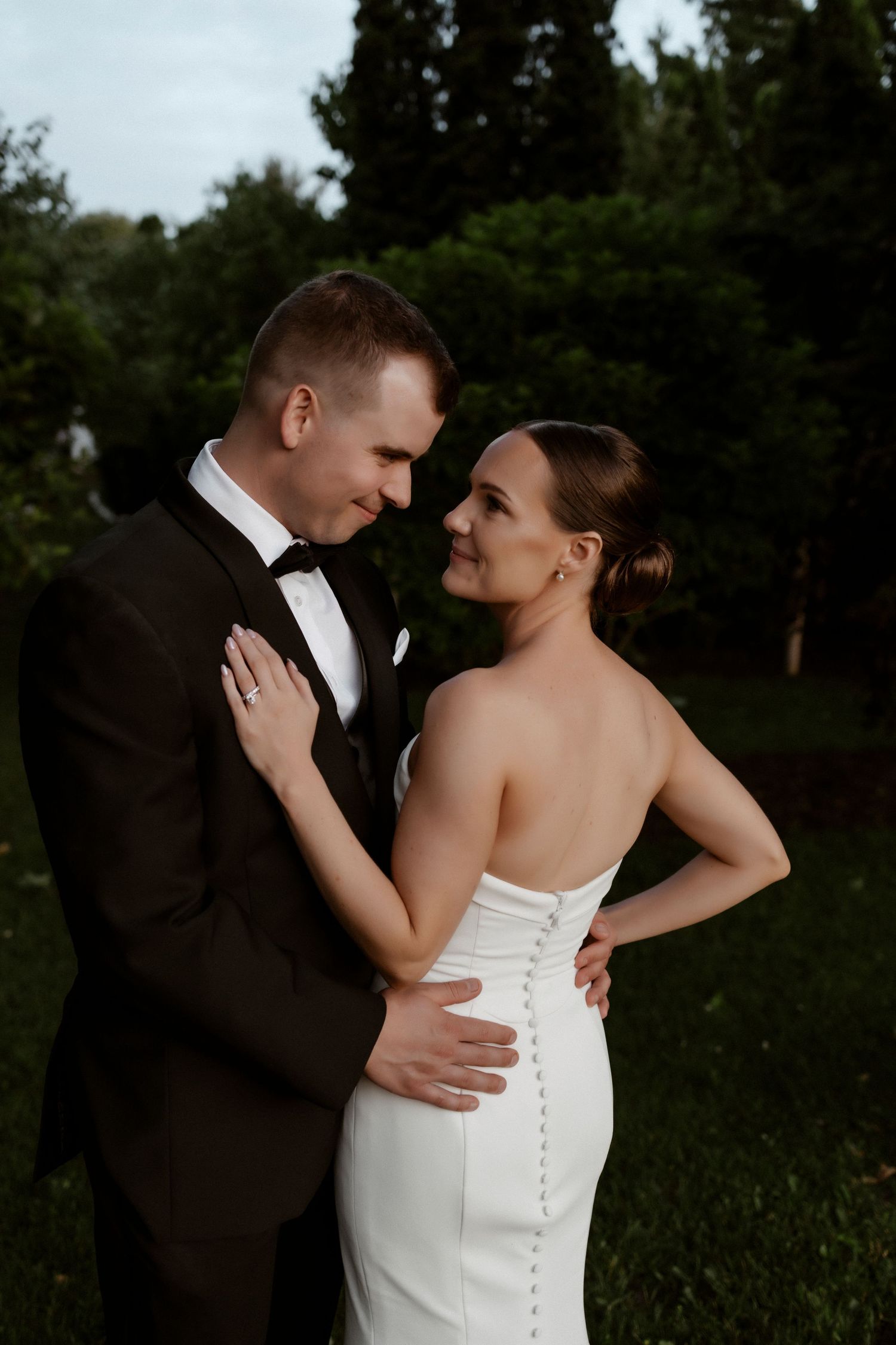 A newlywed couple embraces intimately outdoors, with bride in white gown and groom in black tuxedo at dusk.