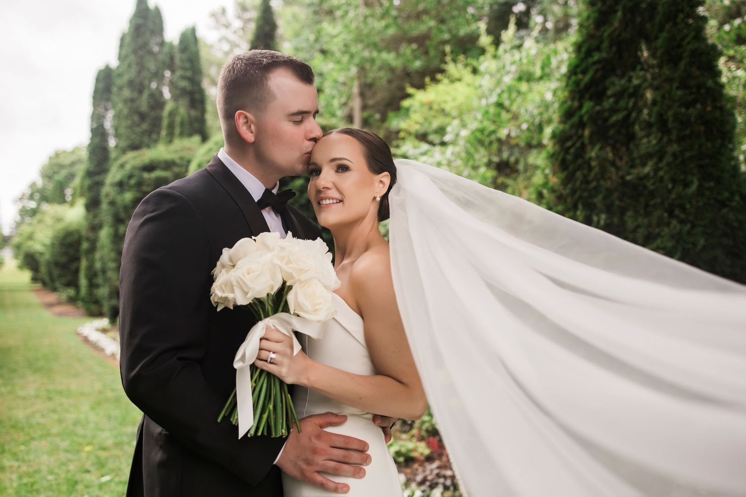 Wedding couple embraces while bride's veil flows elegantly in outdoor garden setting.