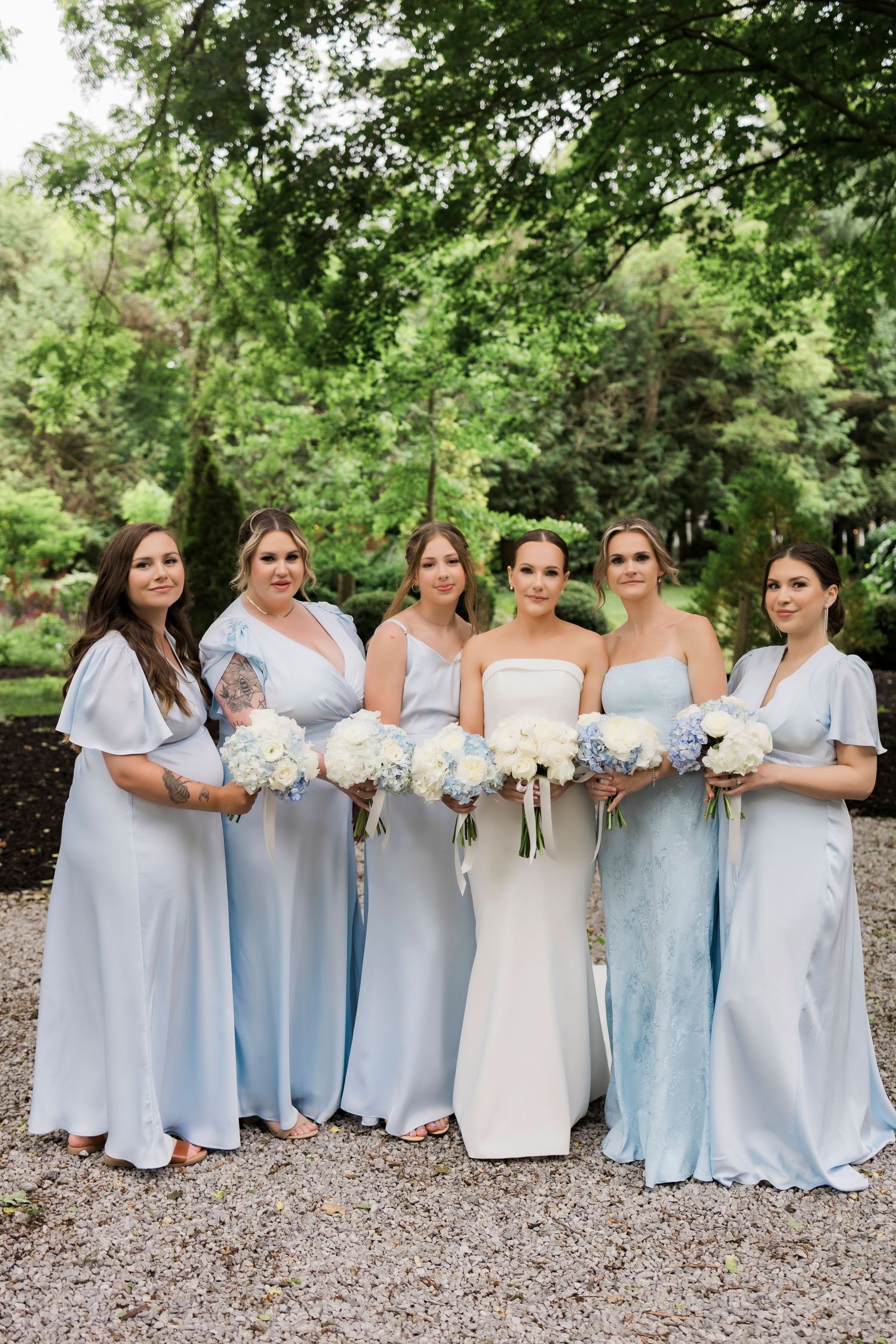 A bridal party in light blue dresses poses together holding white bouquets in a garden setting.