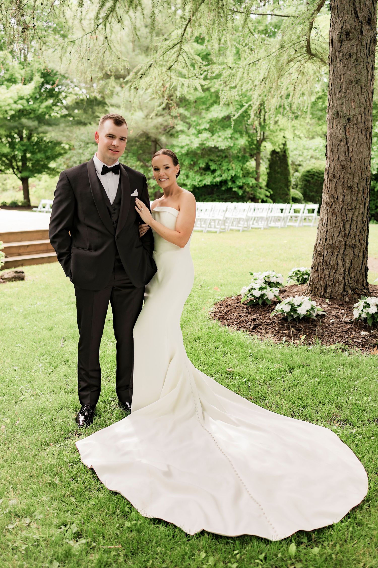 Wedding portrait of couple in formal attire standing under evergreen trees in garden setting.
