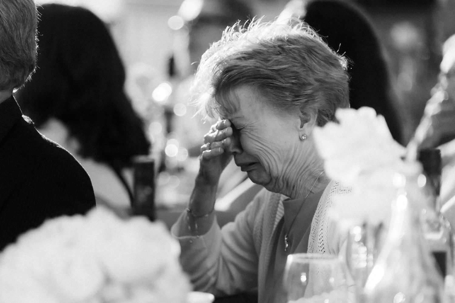 An elegant black and white candid photograph shows a moment of laughter at a dining table with flowers in the foreground.