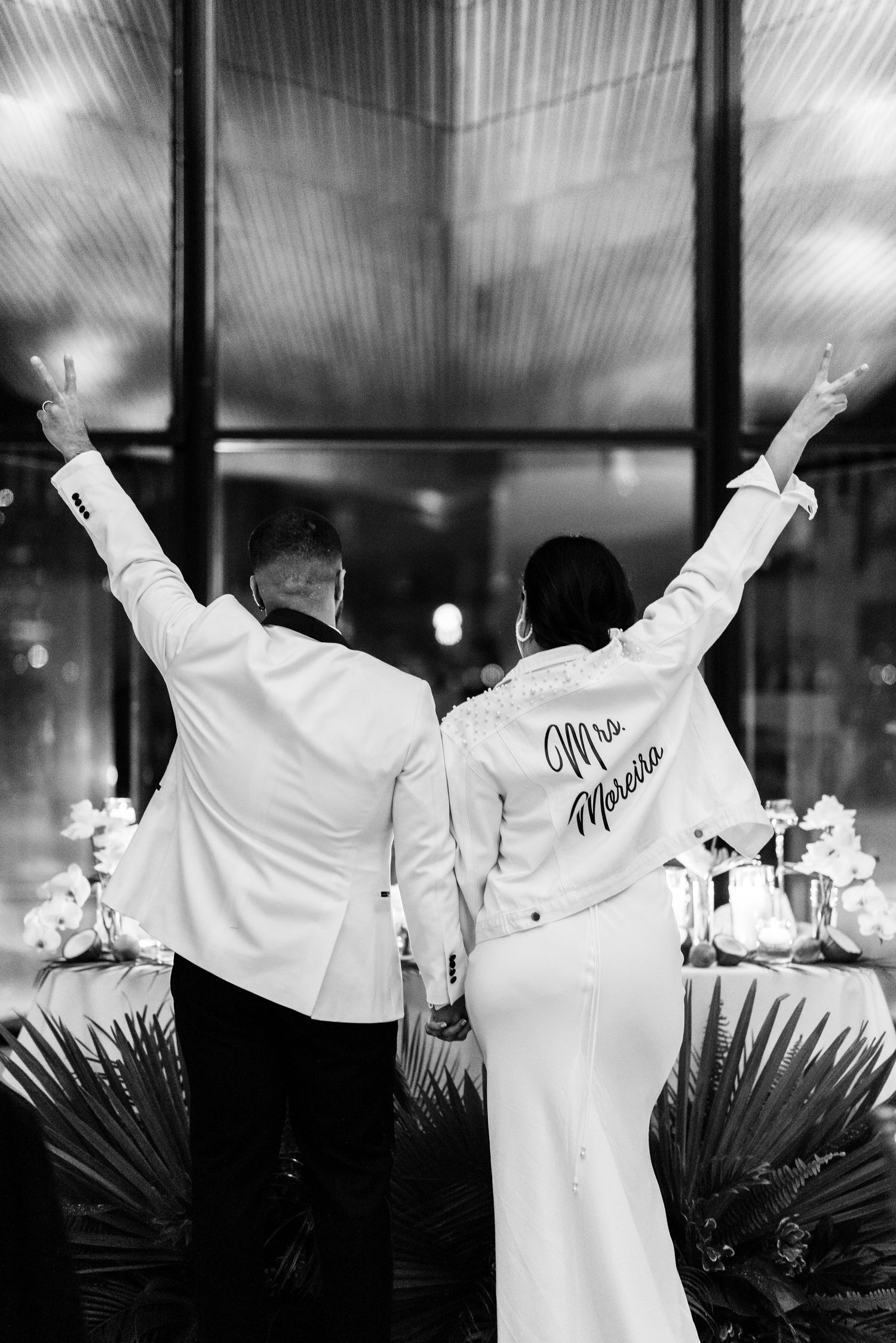Couple in white attire celebrates their wedding with arms raised against a dark window backdrop.