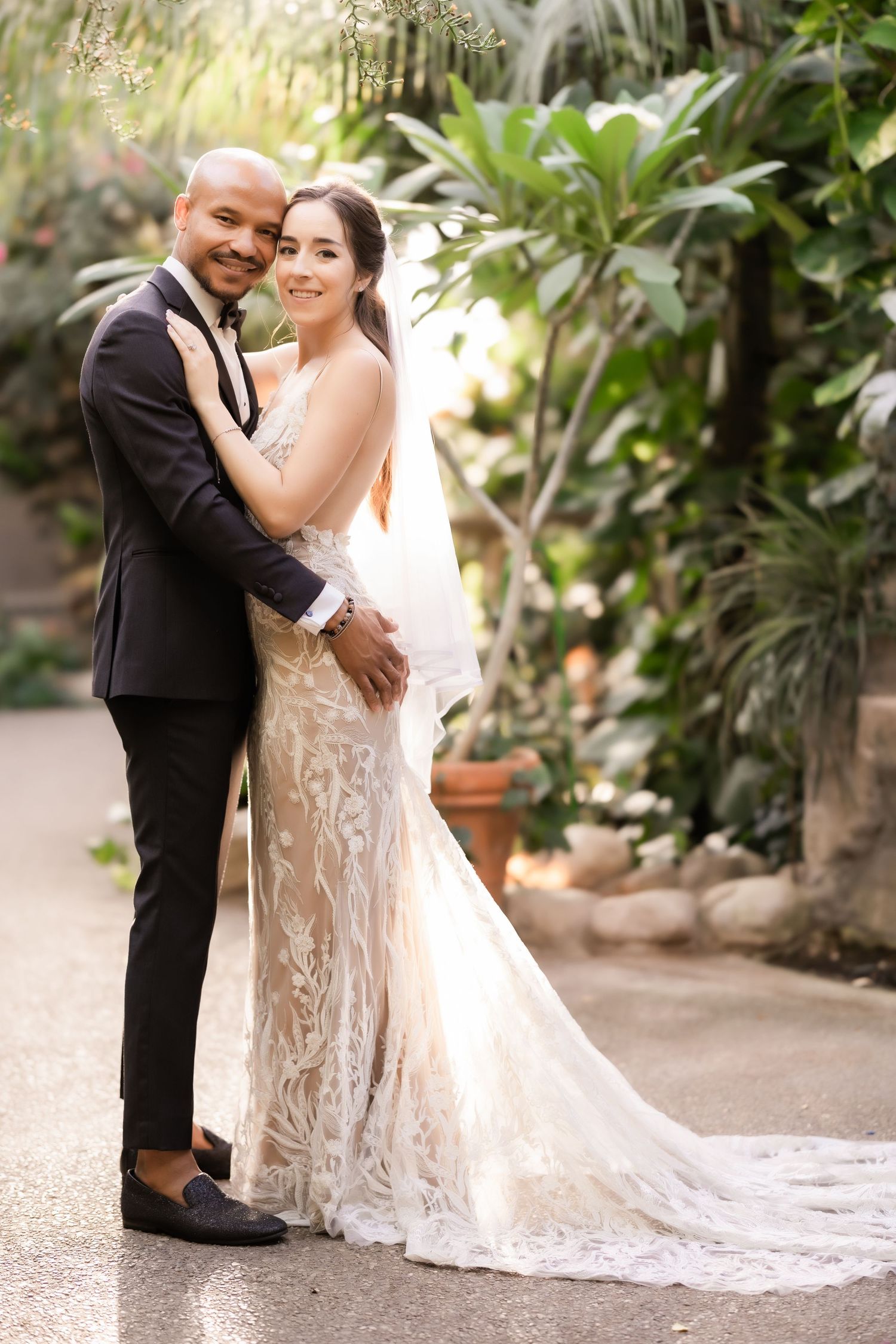 Elegant couple poses in wedding attire against backdrop of tropical plants.