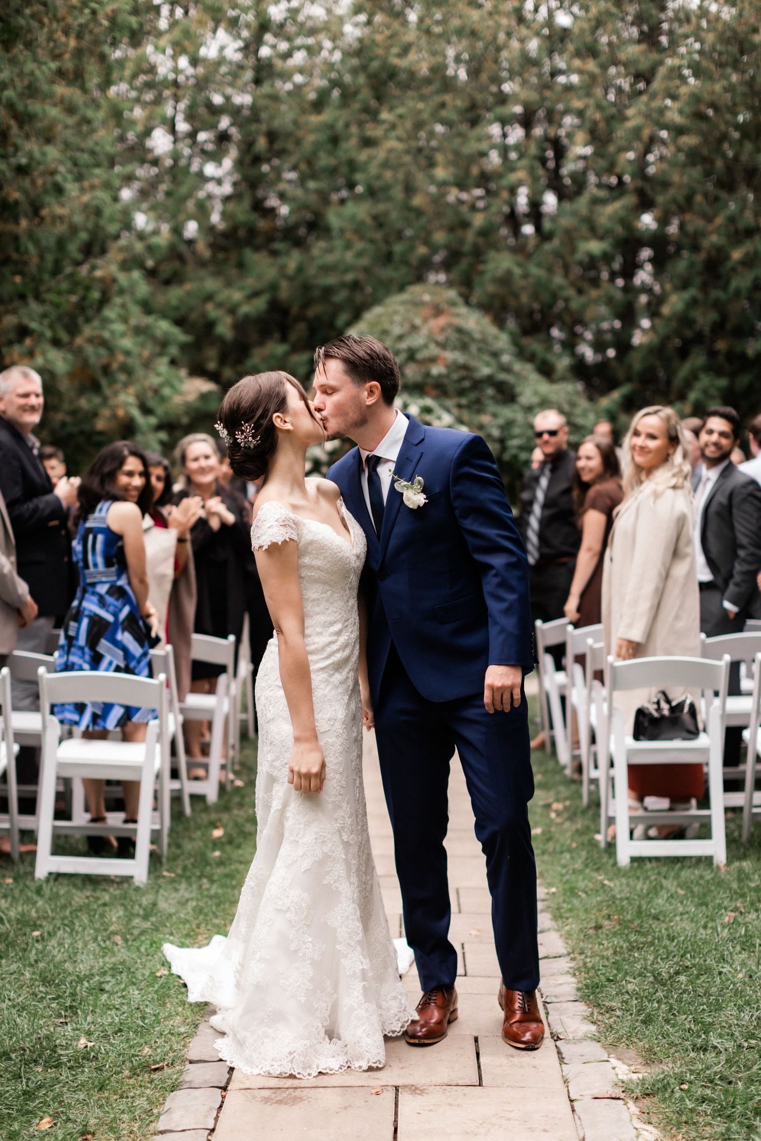 A wedding couple sharing a tender moment on a stone pathway with guests and white chairs in the background.