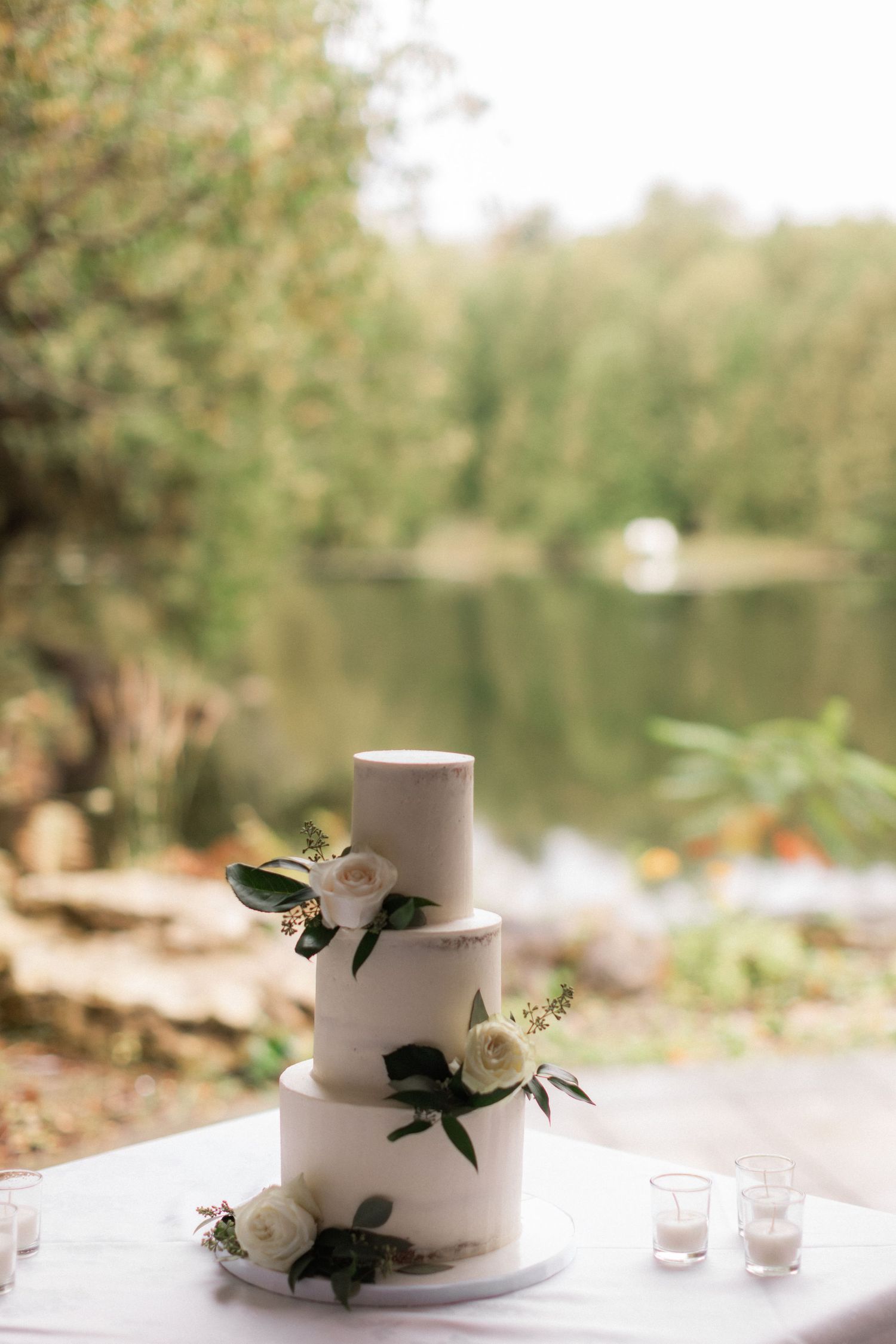 Minimalist white wedding cake with fresh flowers against lakeside backdrop.