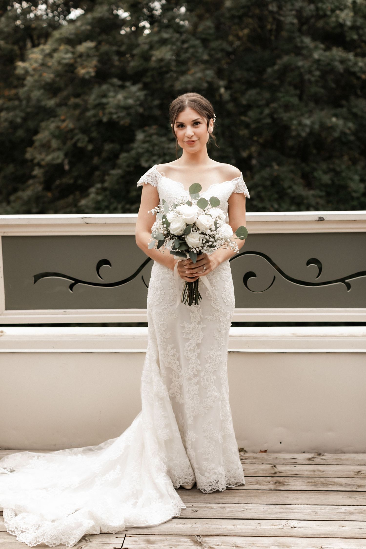 A bride in a lace cap-sleeve gown poses with white bouquet on a balcony.