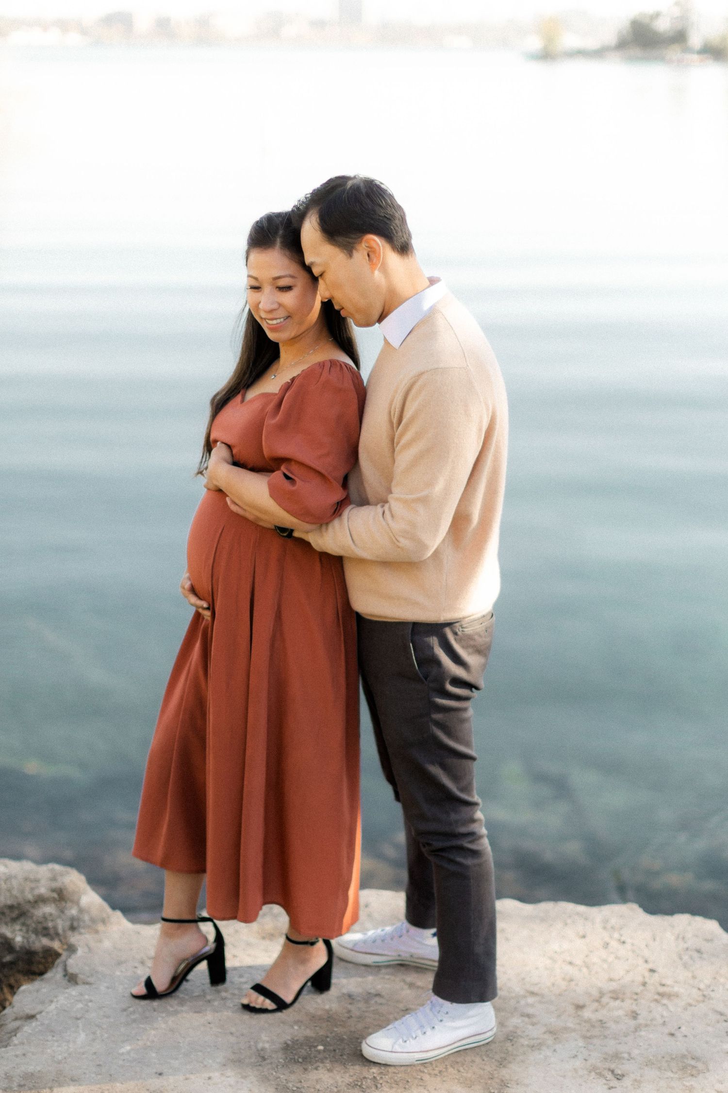 A romantic cliffside maternity photo with a couple in elegant attire overlooking water.