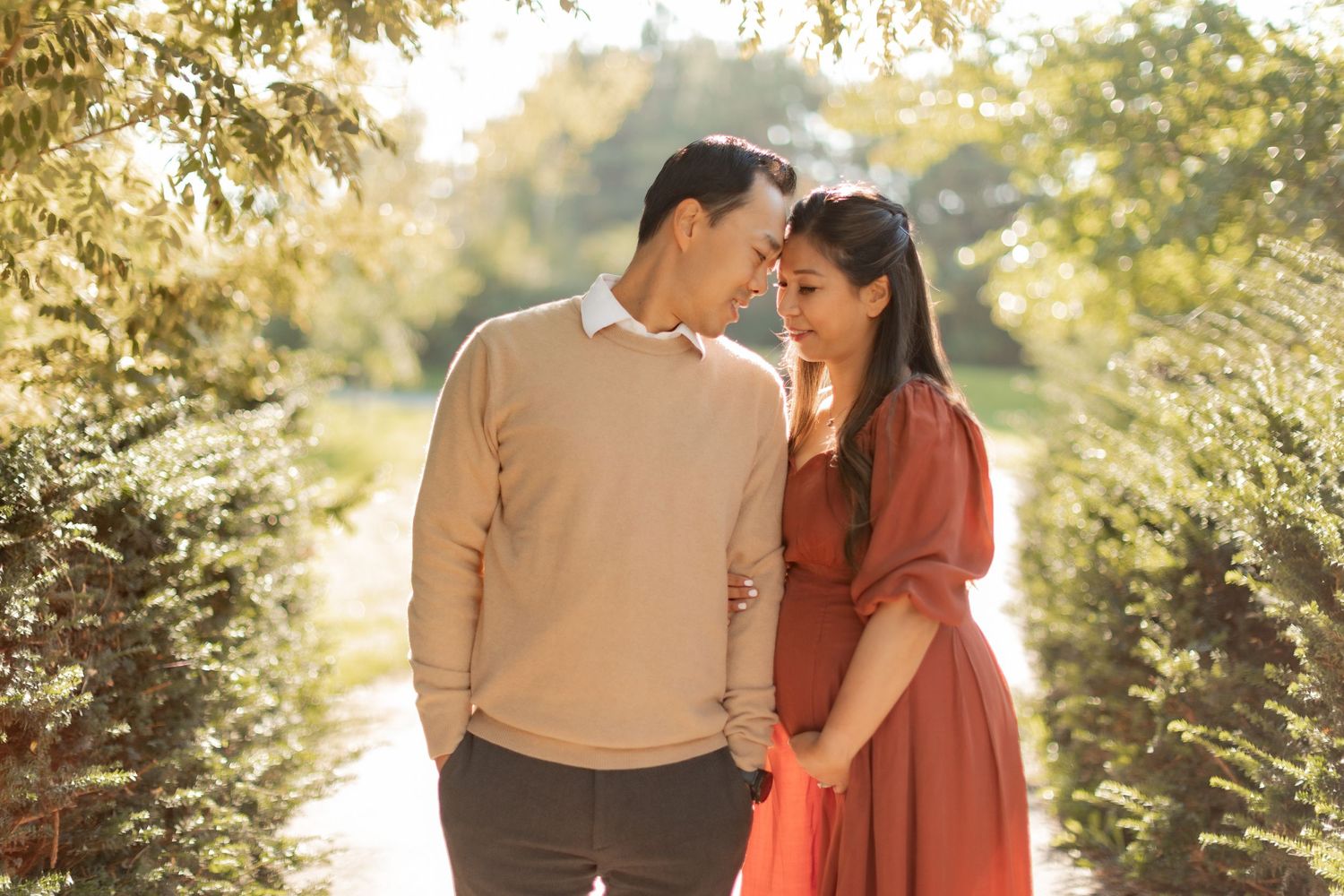 Couple in autumn sunlight sharing an intimate moment among trees, wearing beige sweater and rust dress.