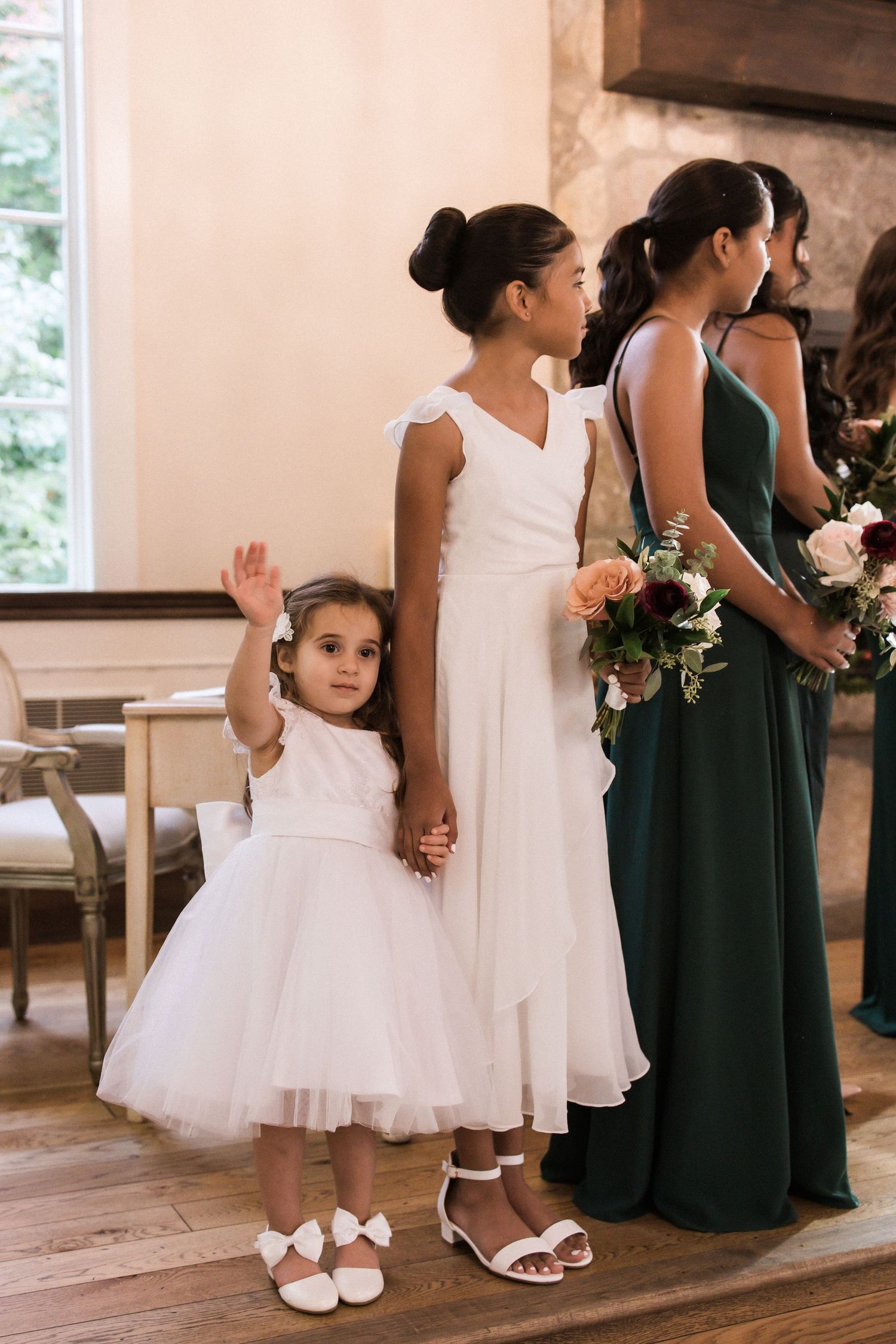 Young flower girl in white dress waves while standing with bridesmaids in elegant venue.