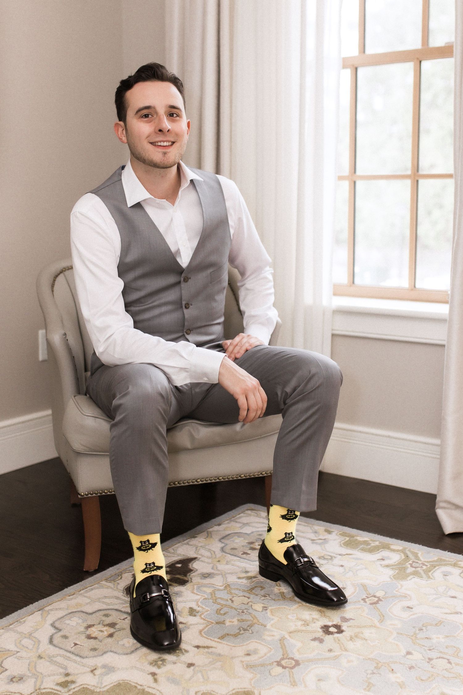 Groom sits in armchair showing off yellow patterned socks with formal grey attire.