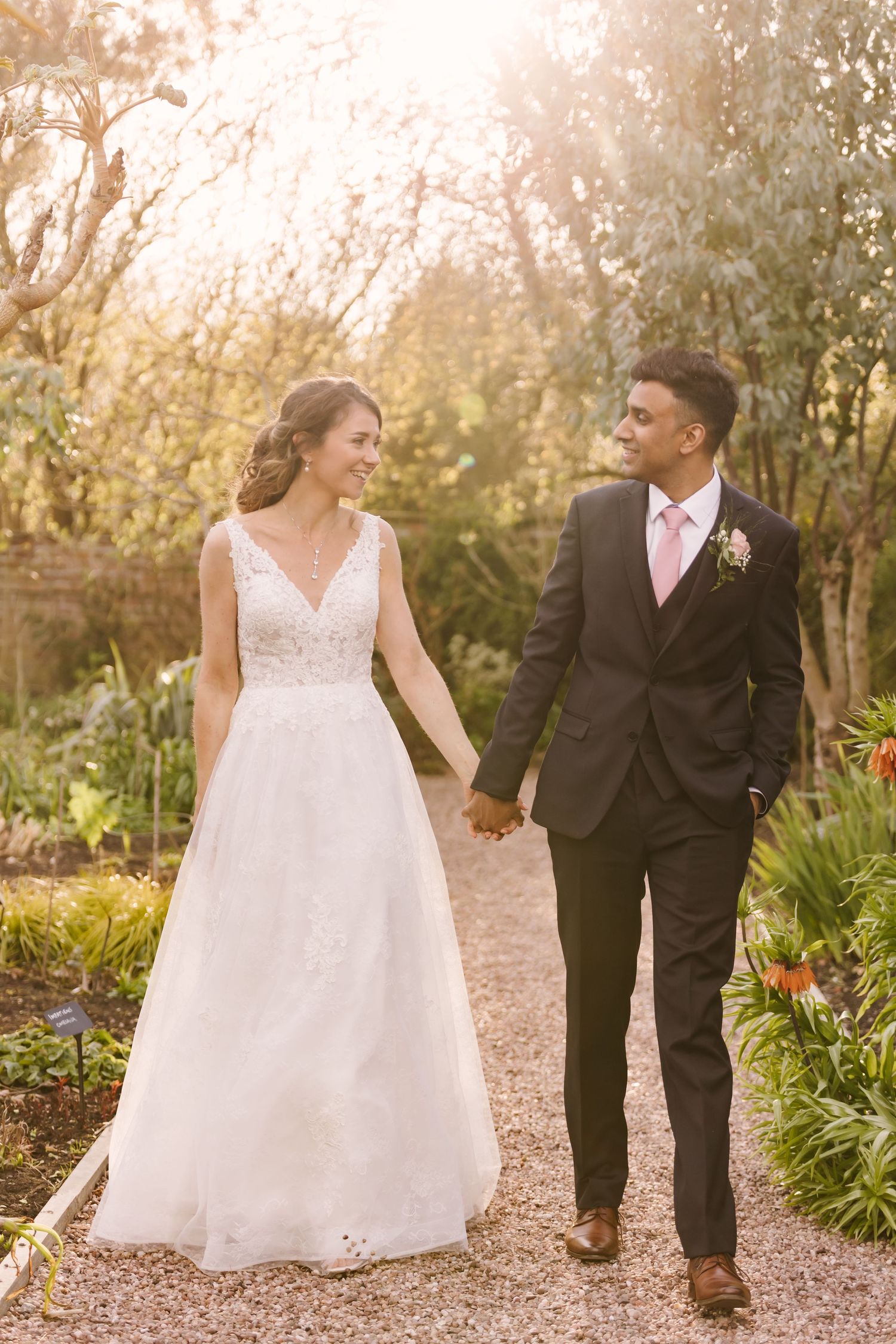 Couple walking hand in hand on garden path in golden sunlight wearing wedding attire.