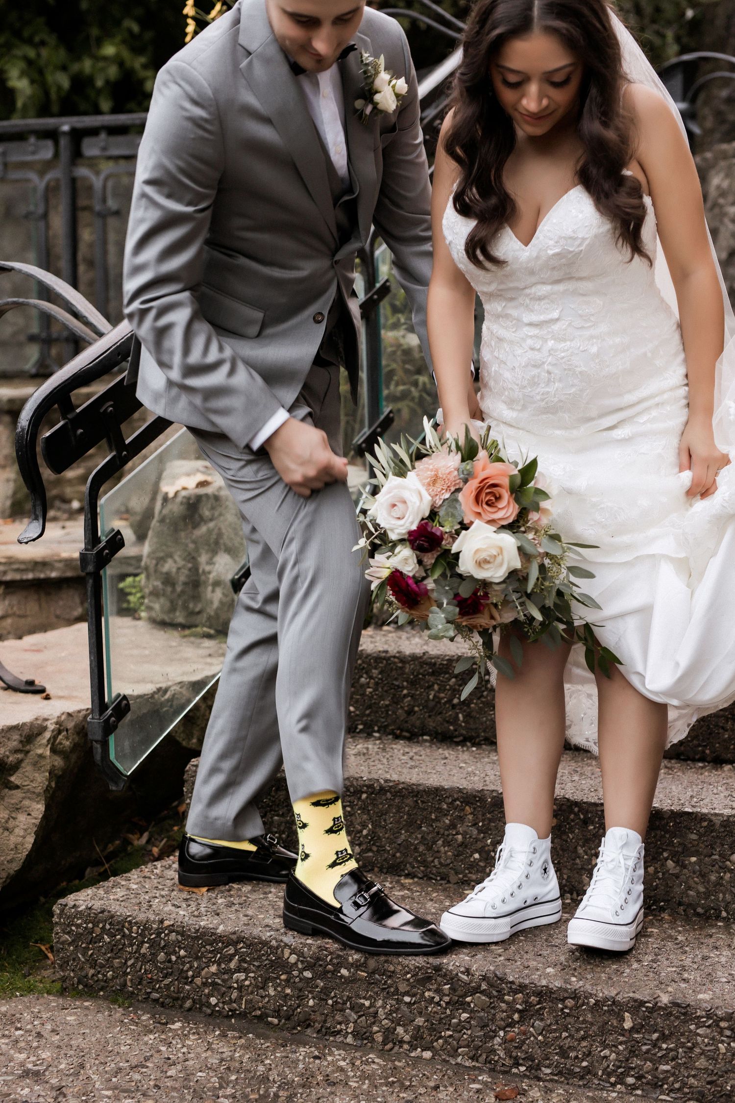 Wedding couple shows off their contrasting footwear while holding a beautiful floral bouquet on stone steps.