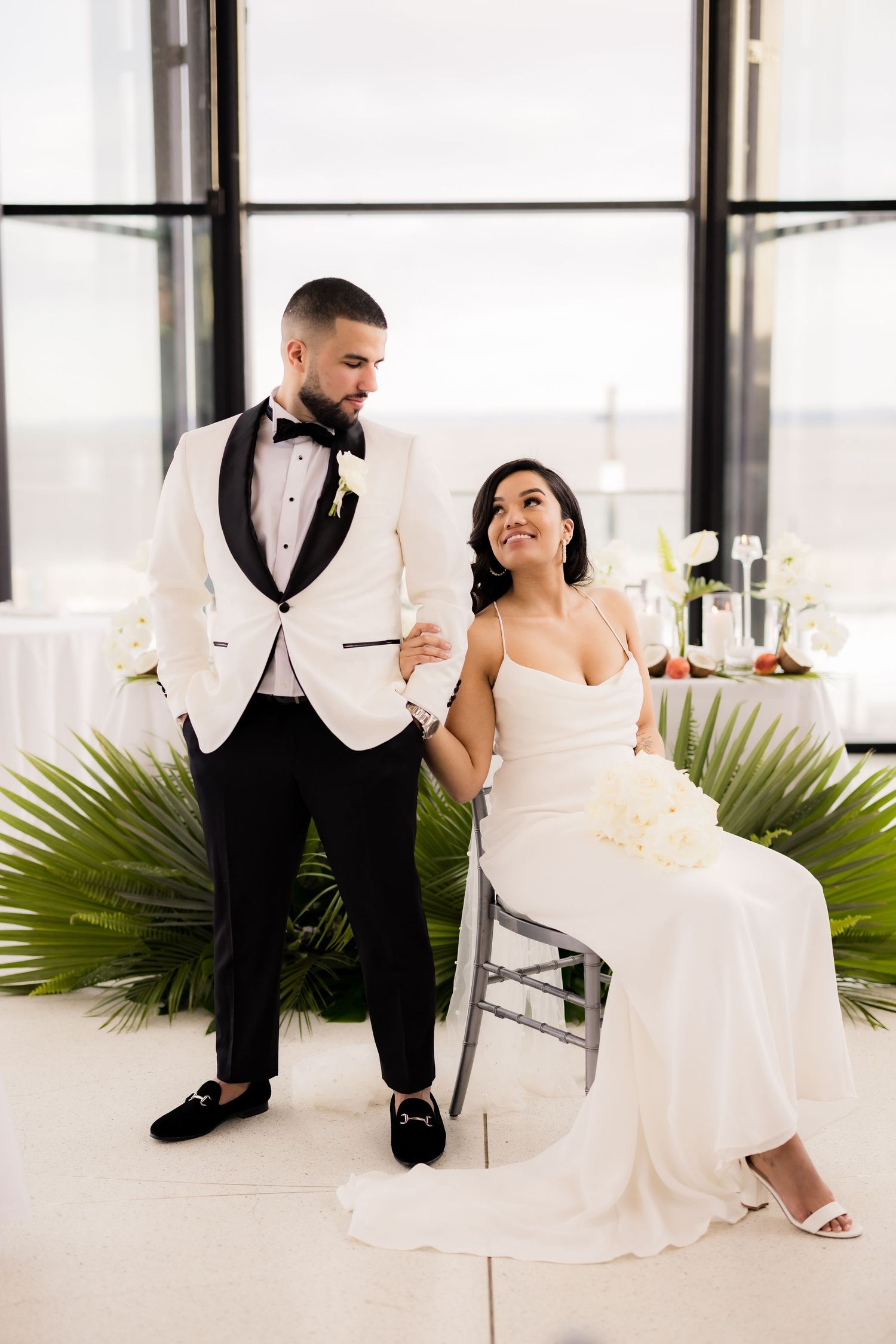 A couple poses at their wedding, with the groom in a white tuxedo and bride in an elegant white gown near palm decor.