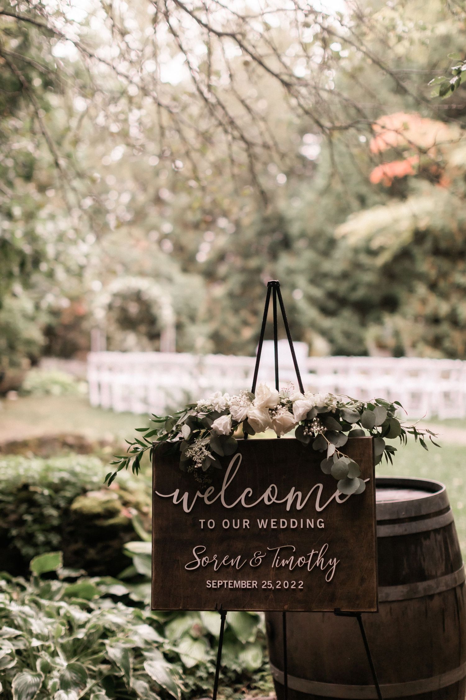 Elegant wooden welcome sign with floral decor displayed on barrel at outdoor wedding venue.