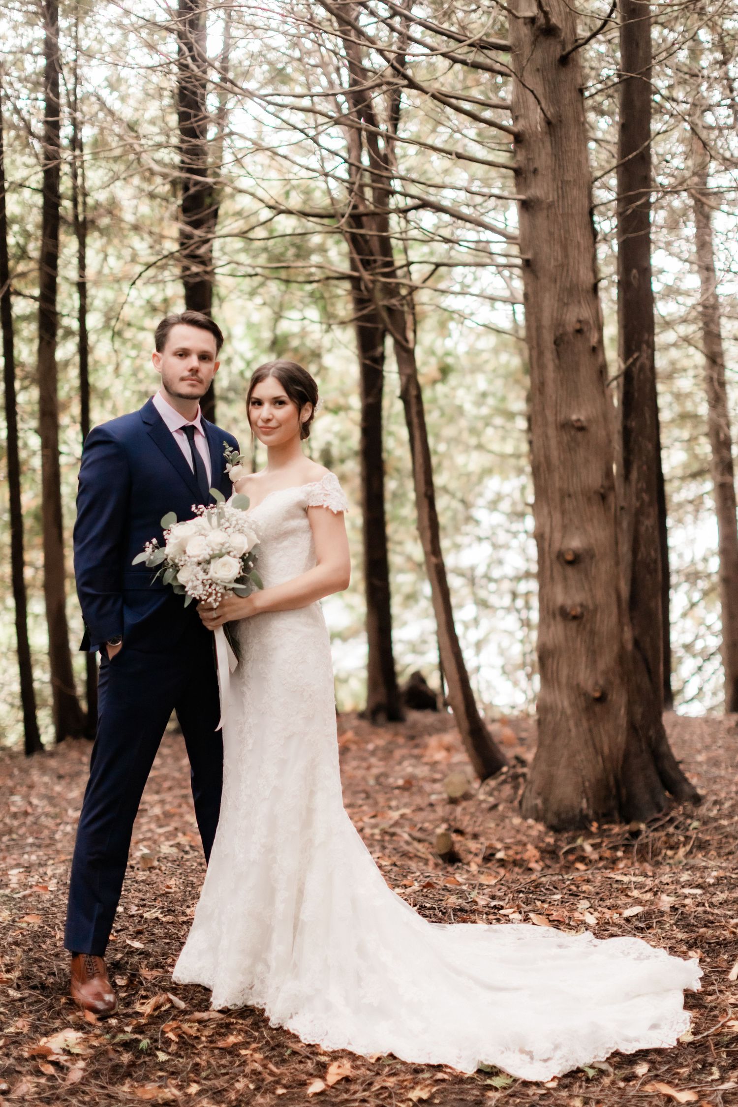 Full-length wedding portrait showing elegant lace dress with train against towering forest trees.