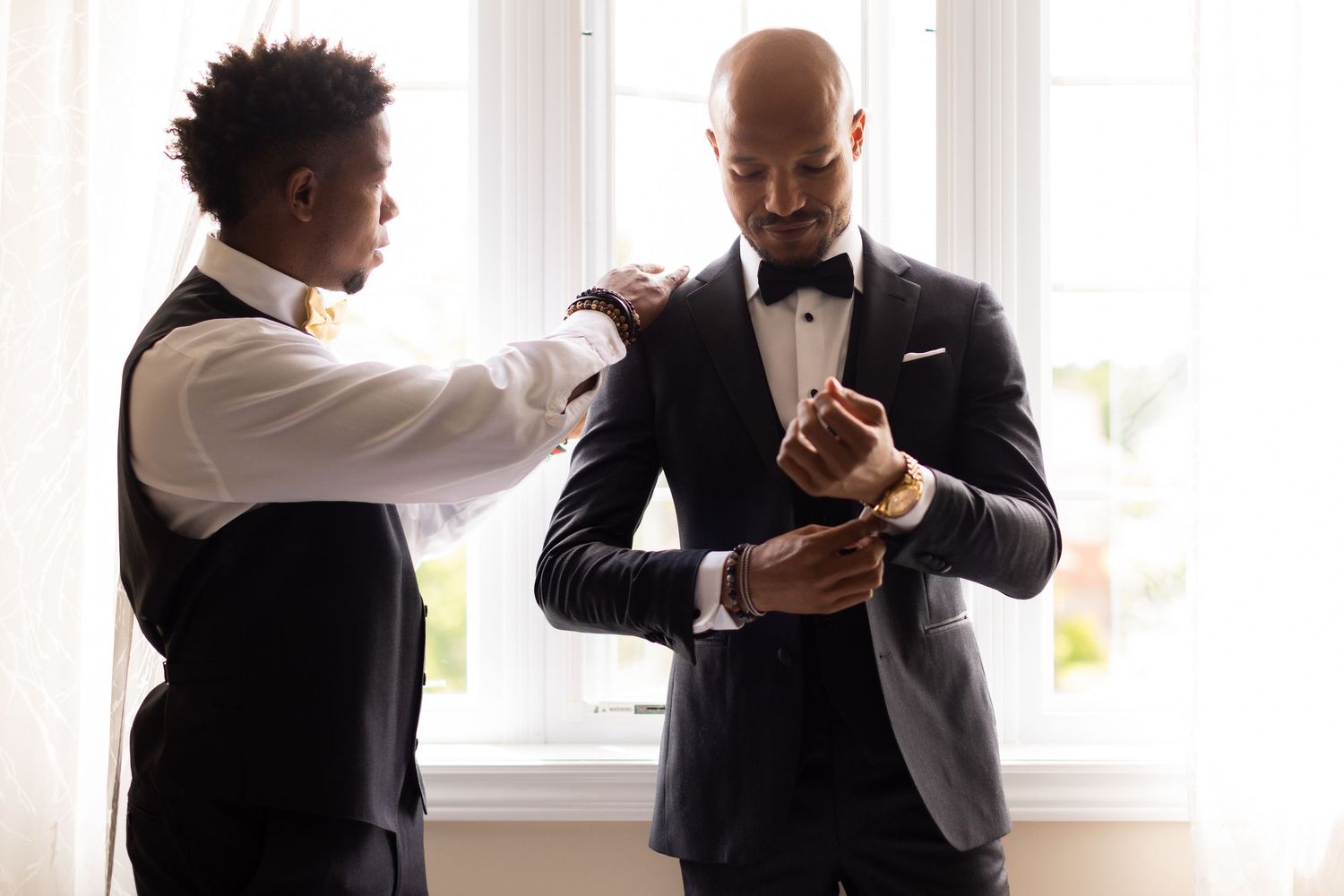 Two people in formal wear help each other prepare for an event near a window.