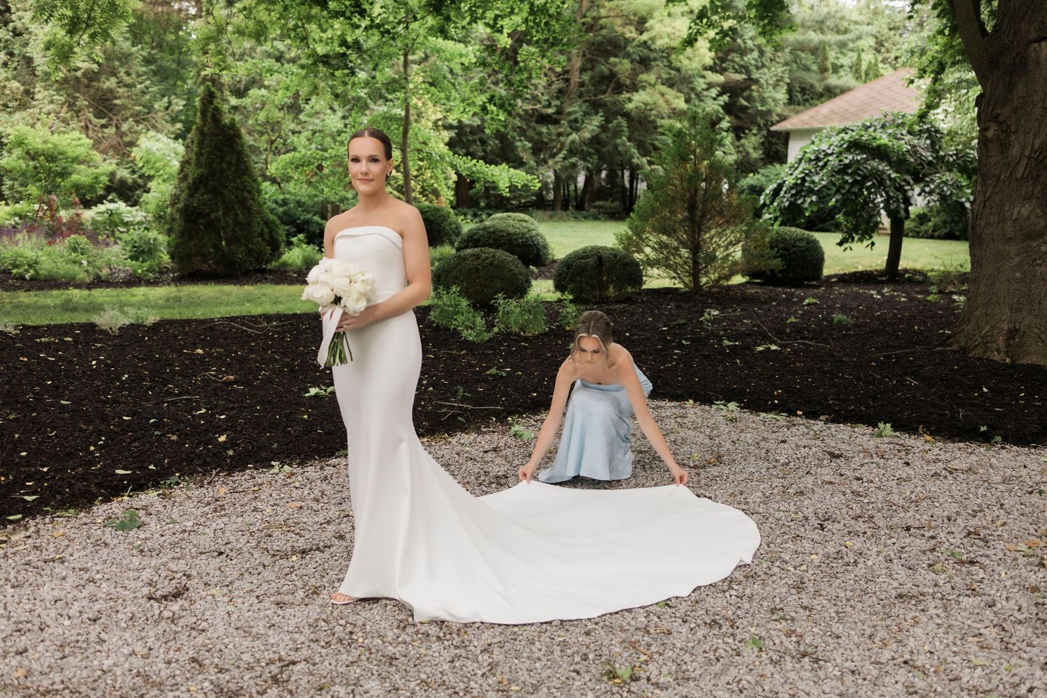 Bride in white wedding dress poses gracefully in a lush garden setting on a gravel path.