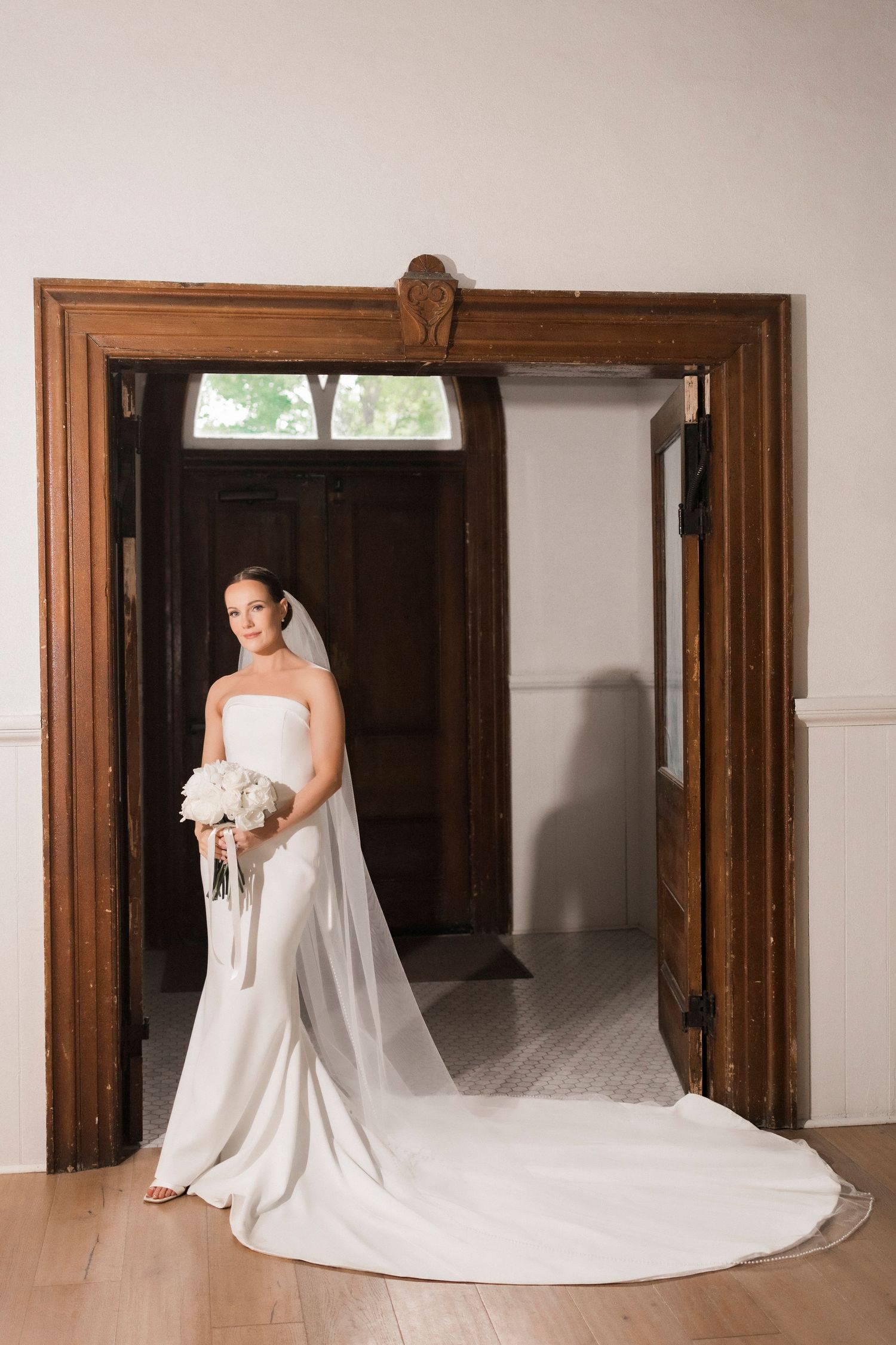 Elegant wedding dress with long train framed by wooden doorway in classic bridal portrait.