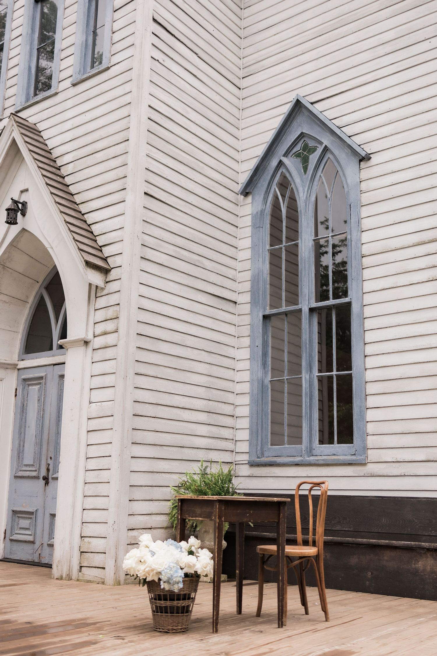White church exterior with gothic window and vintage wooden chair beside floral arrangement.