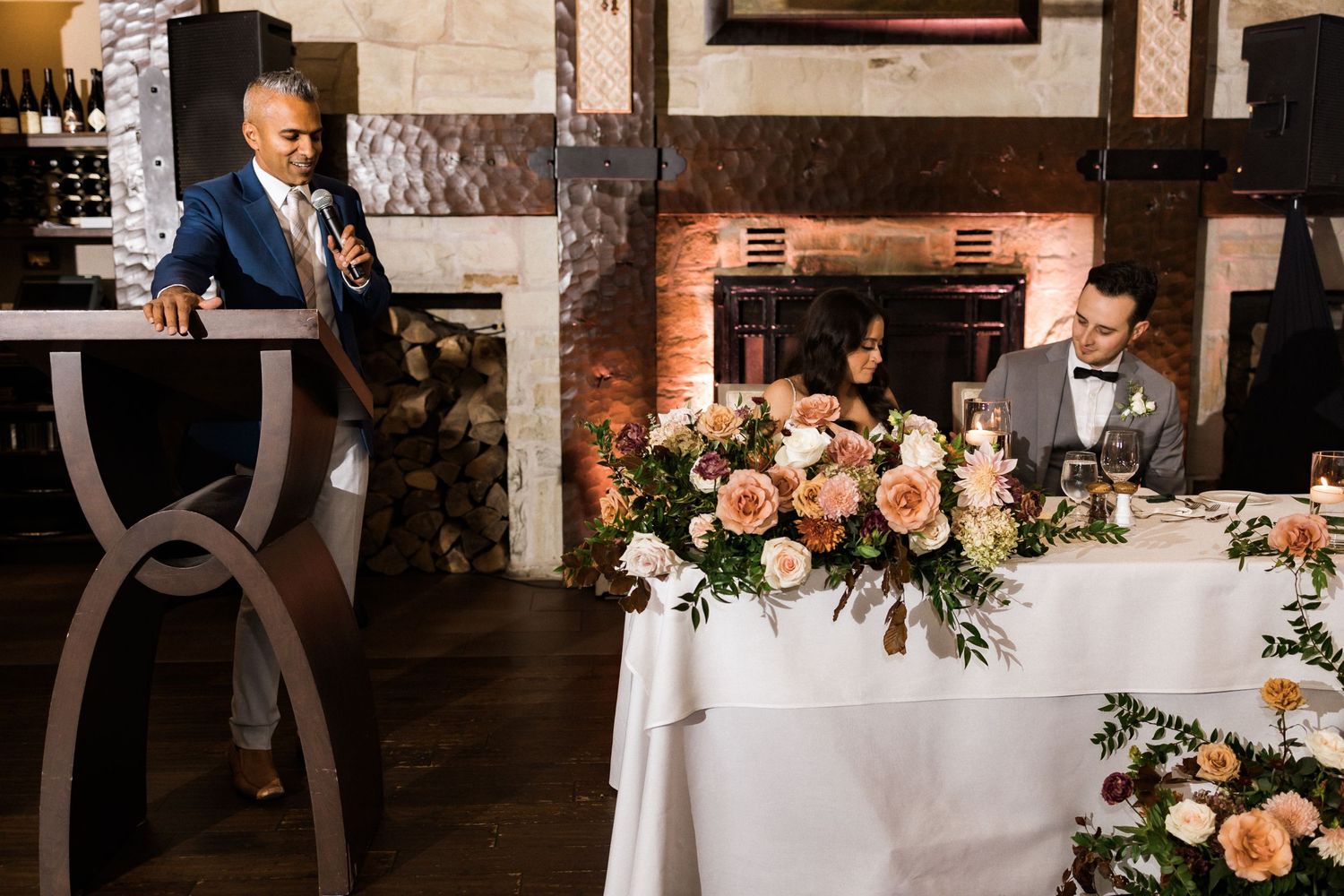 Wedding guests give heartfelt speeches at reception while beautiful floral centerpieces adorn white-clothed tables.
