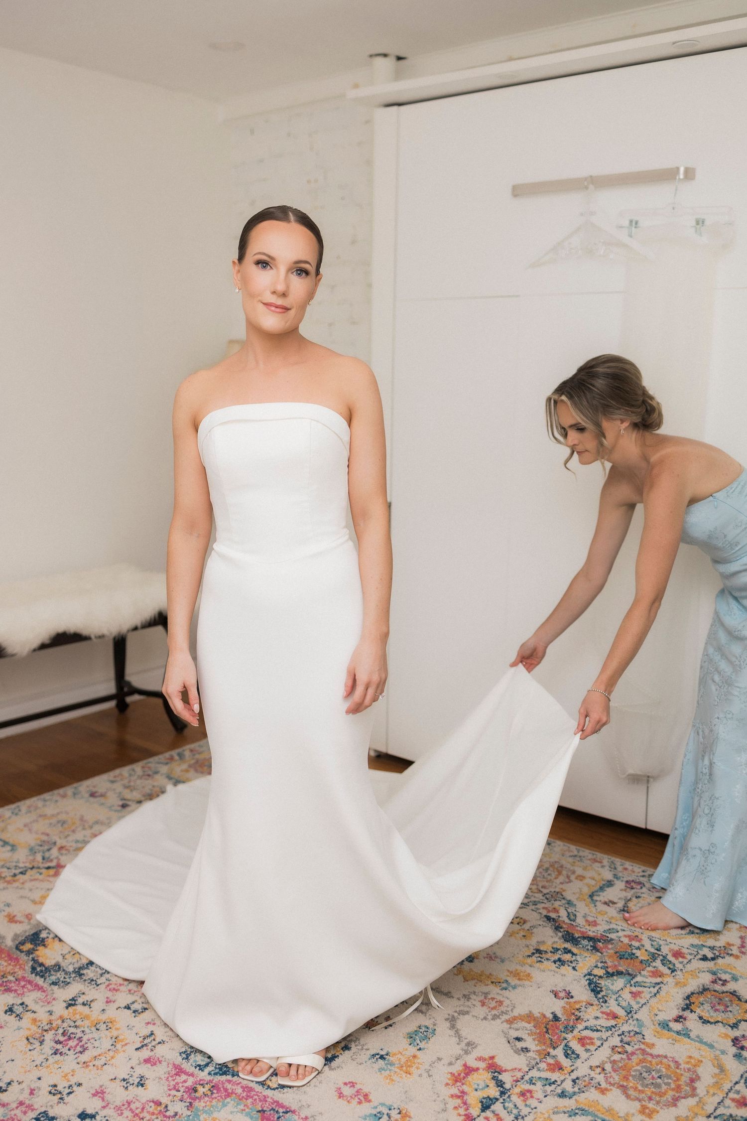 An elegant white wedding dress being adjusted on a decorative rug in a bright interior room.