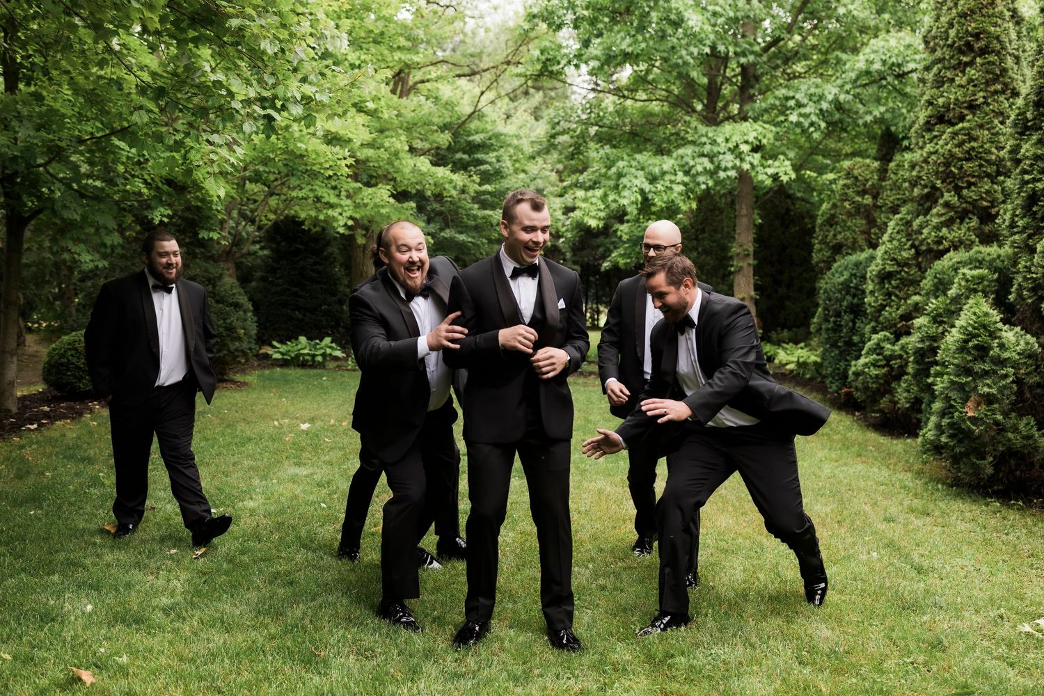 Wedding party in black suits walks playfully across green lawn during outdoor celebration.