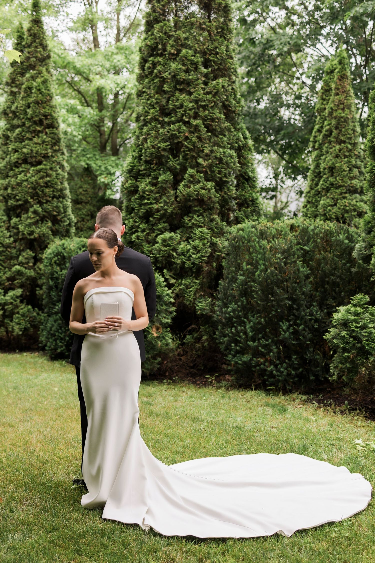 Romantic wedding couple embraces in a garden setting with flowing white dress train on green lawn.
