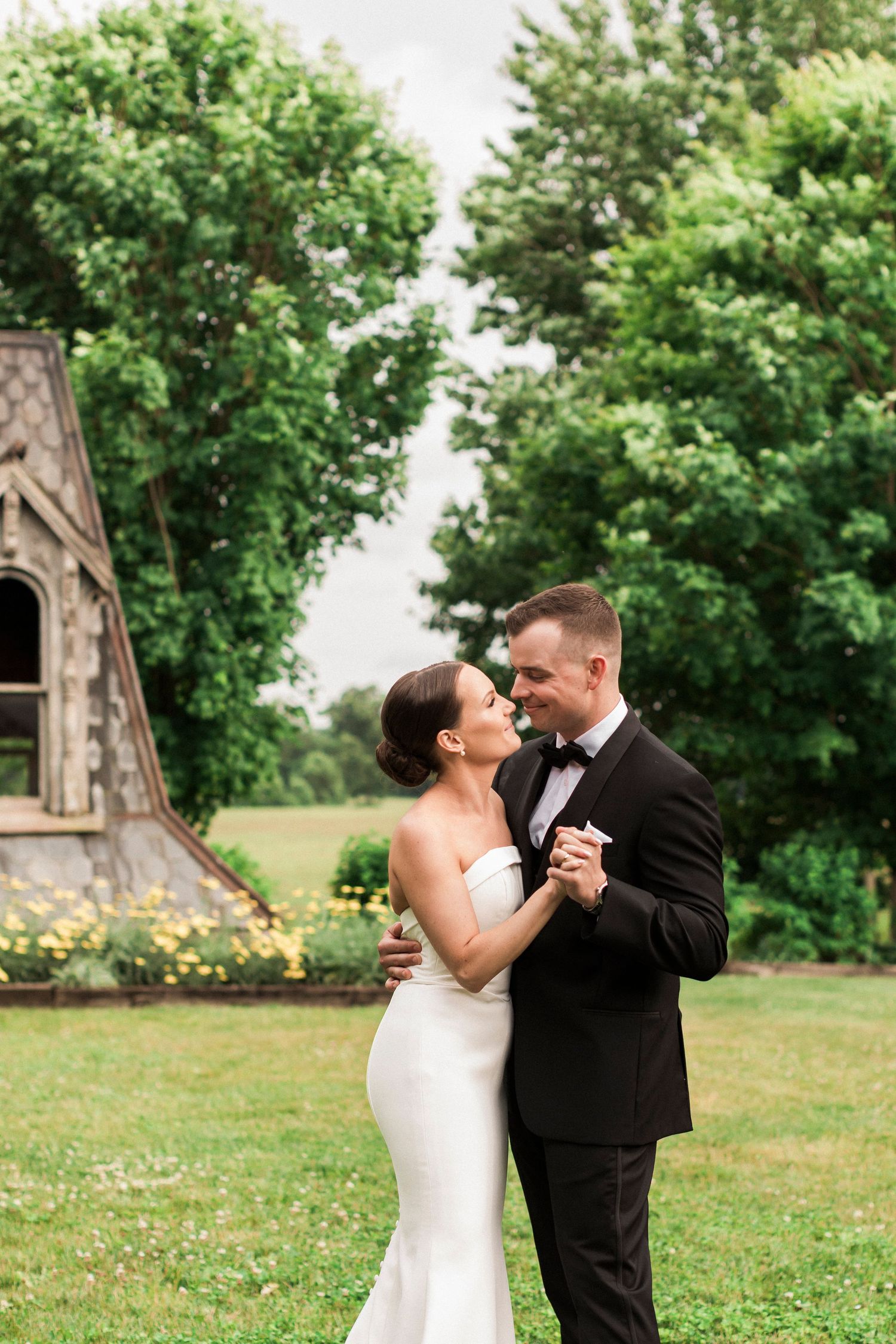 A romantic couple shares their first dance on a beautiful lawn with a rustic chapel in the background.