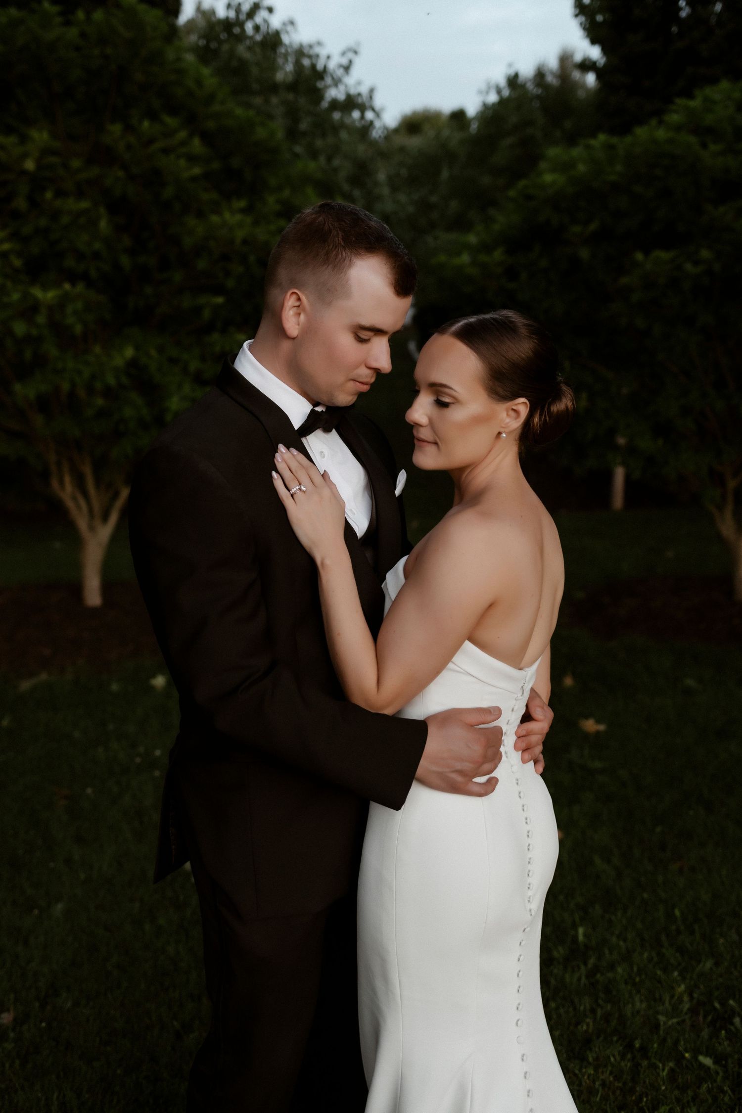 A romantic wedding couple embraces in an intimate outdoor garden setting at dusk.