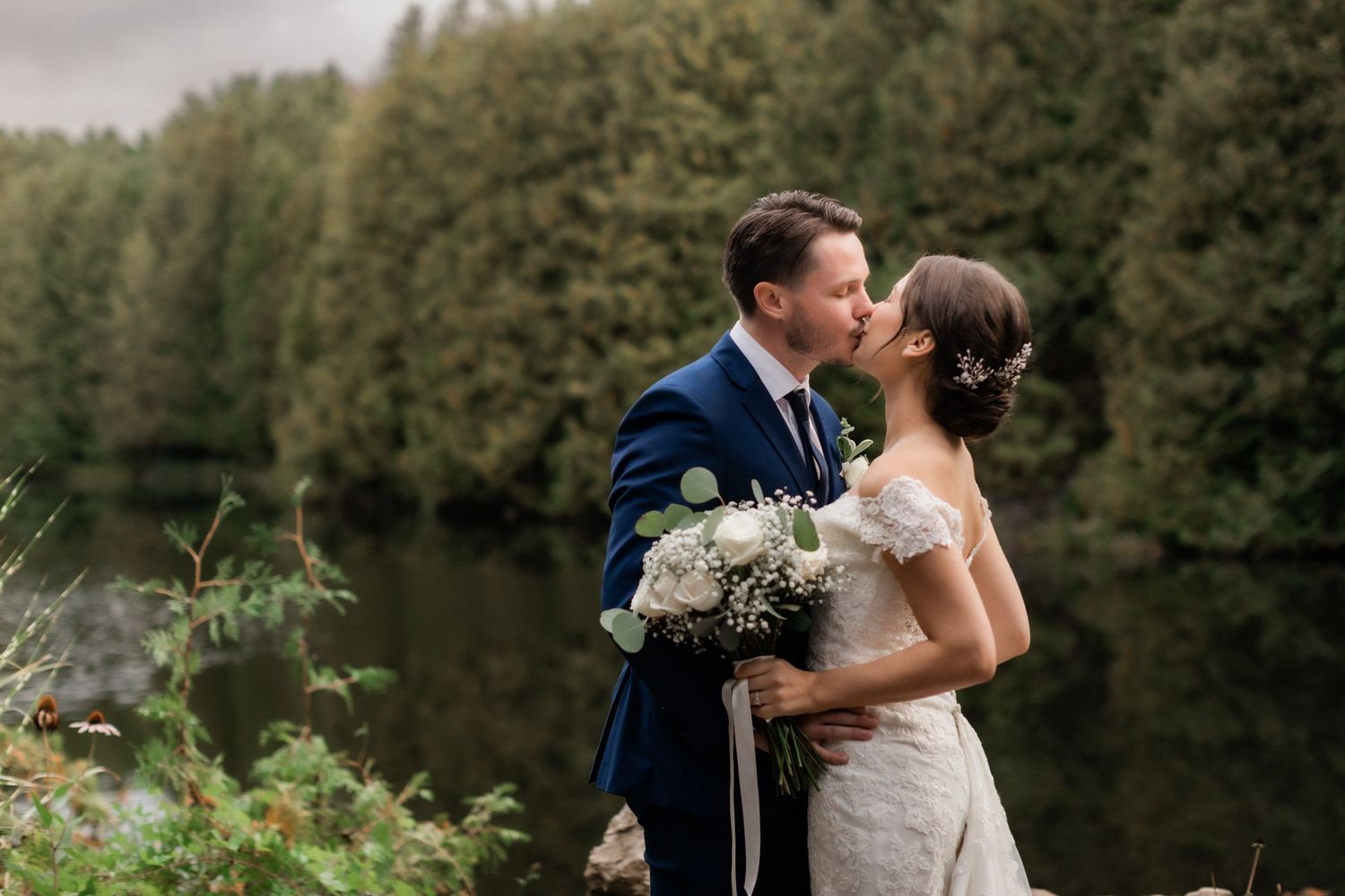 Romantic outdoor wedding portrait by a serene lake with lush forest backdrop.