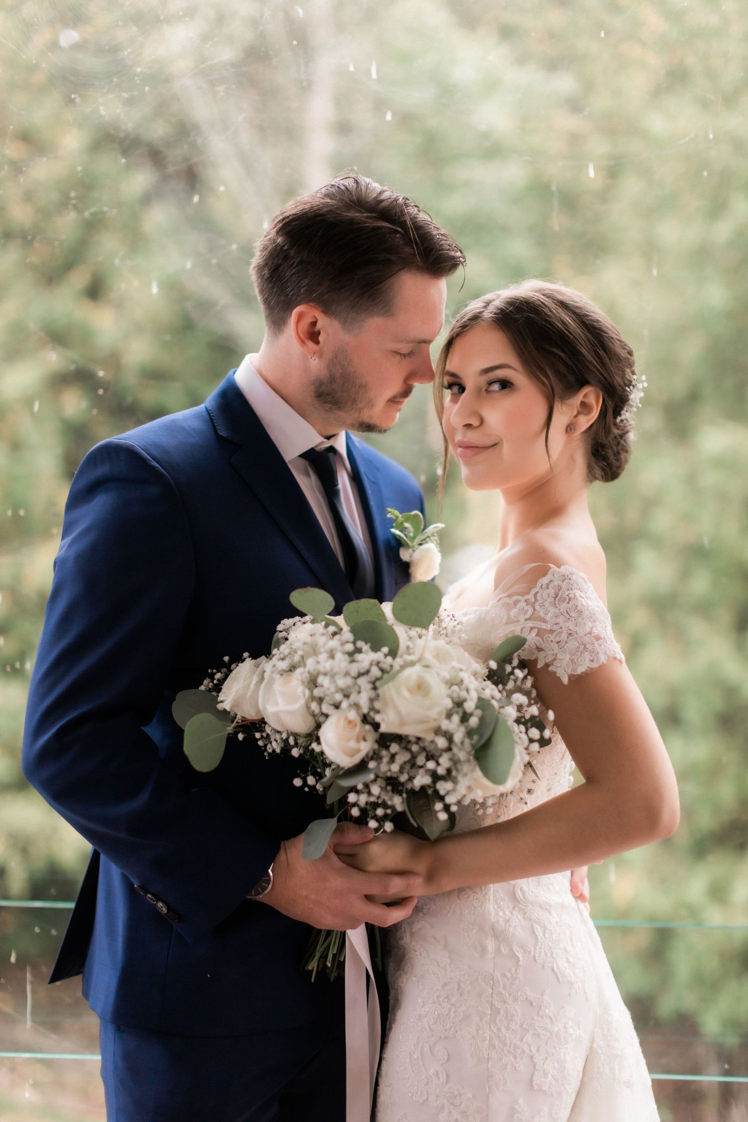 Romantic portrait of newlyweds with white floral bouquet against natural outdoor backdrop.