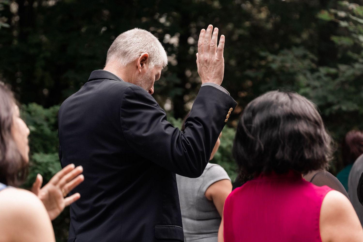 Wedding guest raises hand in celebration during outdoor ceremony amongst trees.