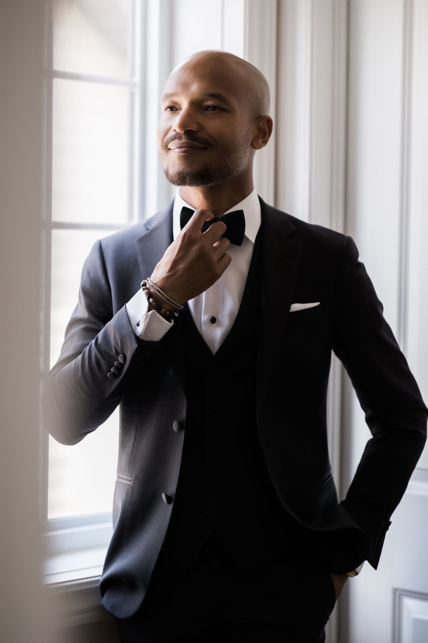 Groom adjusts bow tie while standing near window in elegant three-piece tuxedo.
