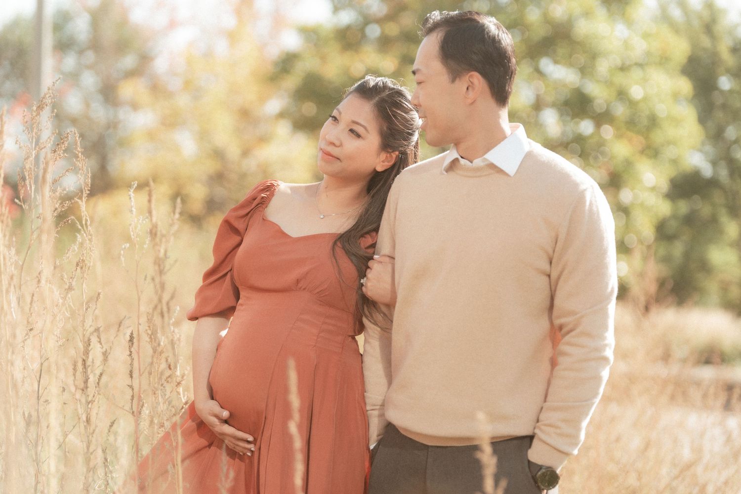 Couple shares intimate moment in golden field as expectant mother in rust-colored dress cradles baby bump.