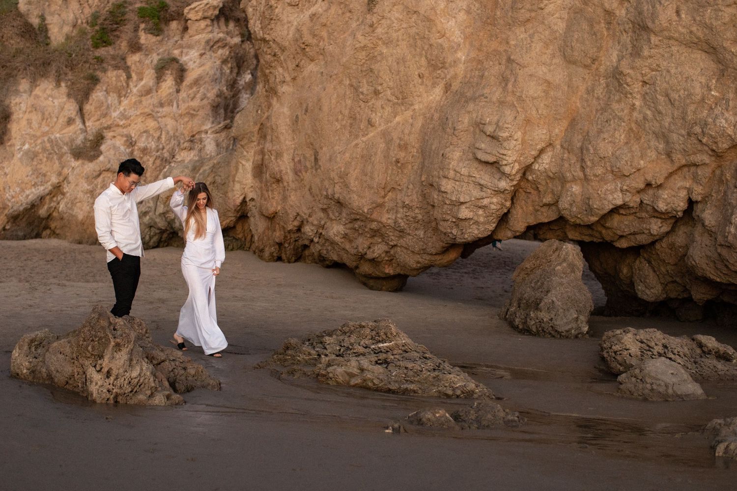 Couple in white outfits standing among coastal rock formations.