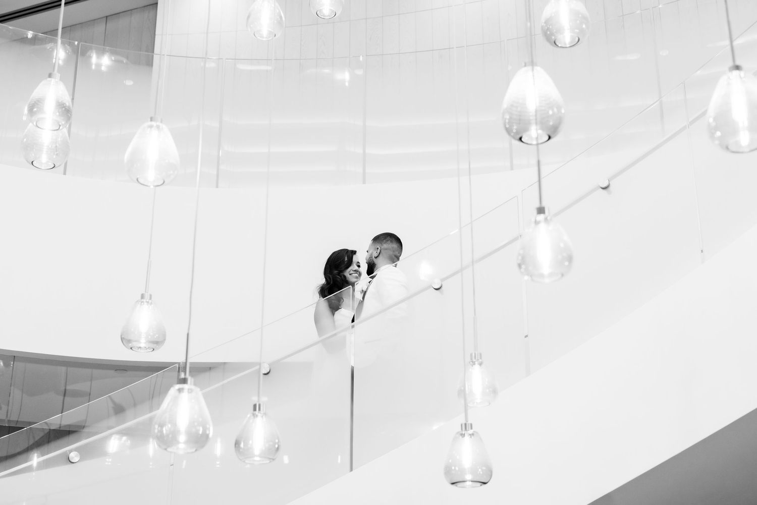 Artistic black and white photo of couple on modern staircase with hanging glass pendant lights.